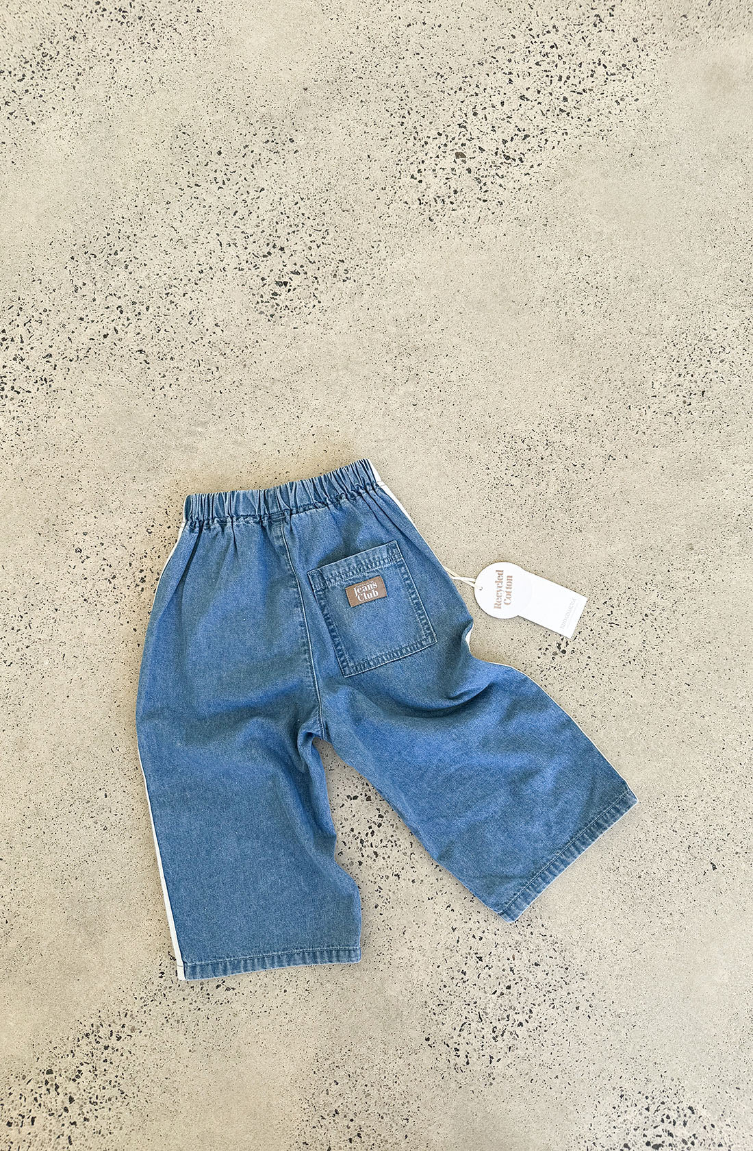 A pair of blue denim Tricky Track Pants by TWIN COLLECTIVE, specifically designed for kids, is laid flat on a speckled concrete surface. Featuring a vintage-inspired design, these pants include an elastic waistband, a back pocket adorned with a small brown patch, and a white tag attached to the waistband.