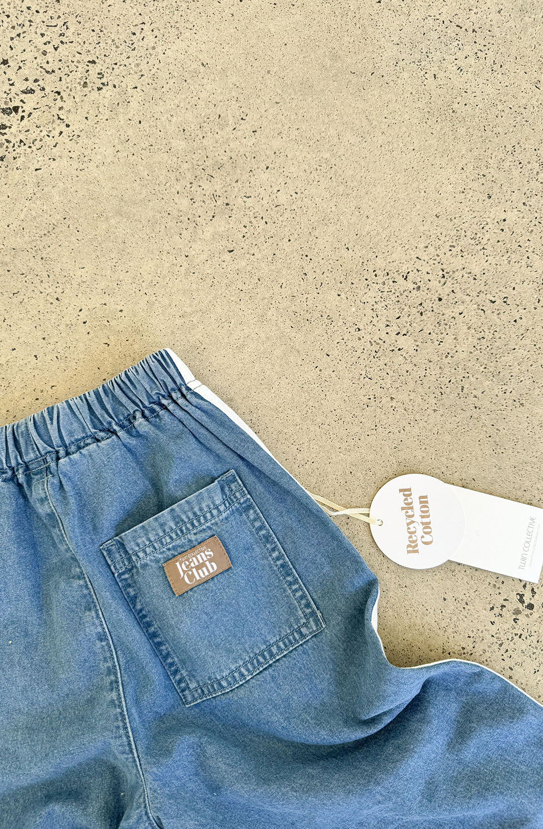 A pair of Tricky Track Pant blue denim jeans with an elastic waistband and a back pocket featuring a brown label that reads "Jeans Club." Attached to the waistband is a white tag labeled "Recycled Cotton." These Kids Tricky Track denim pants by TWIN COLLECTIVE are laid on a textured, speckled surface.