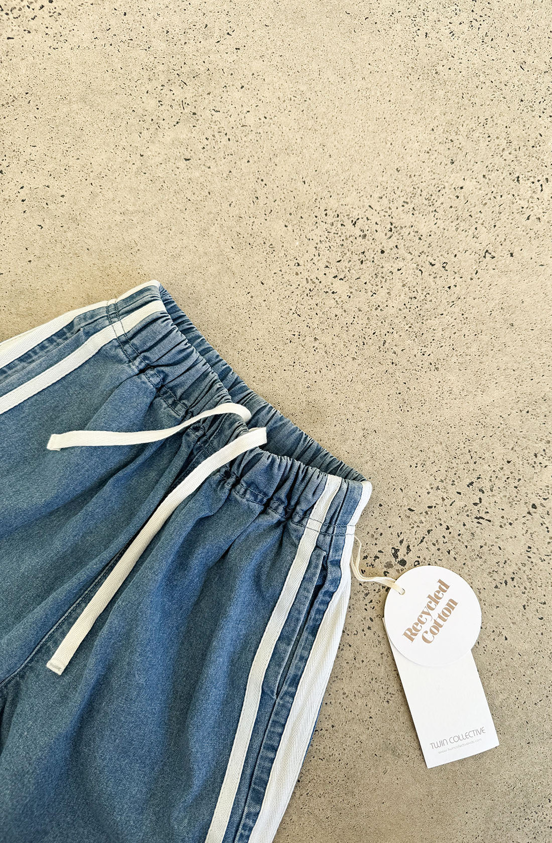 A pair of blue, vintage-inspired sweatpants called the Tricky Track Pant, by TWIN COLLECTIVE. They feature white side stripes and a white drawstring and are displayed on a speckled beige surface. The oversized track pants have a tag attached with text that reads "Recycled Cotton" and "TWIN COLLECTIVE.