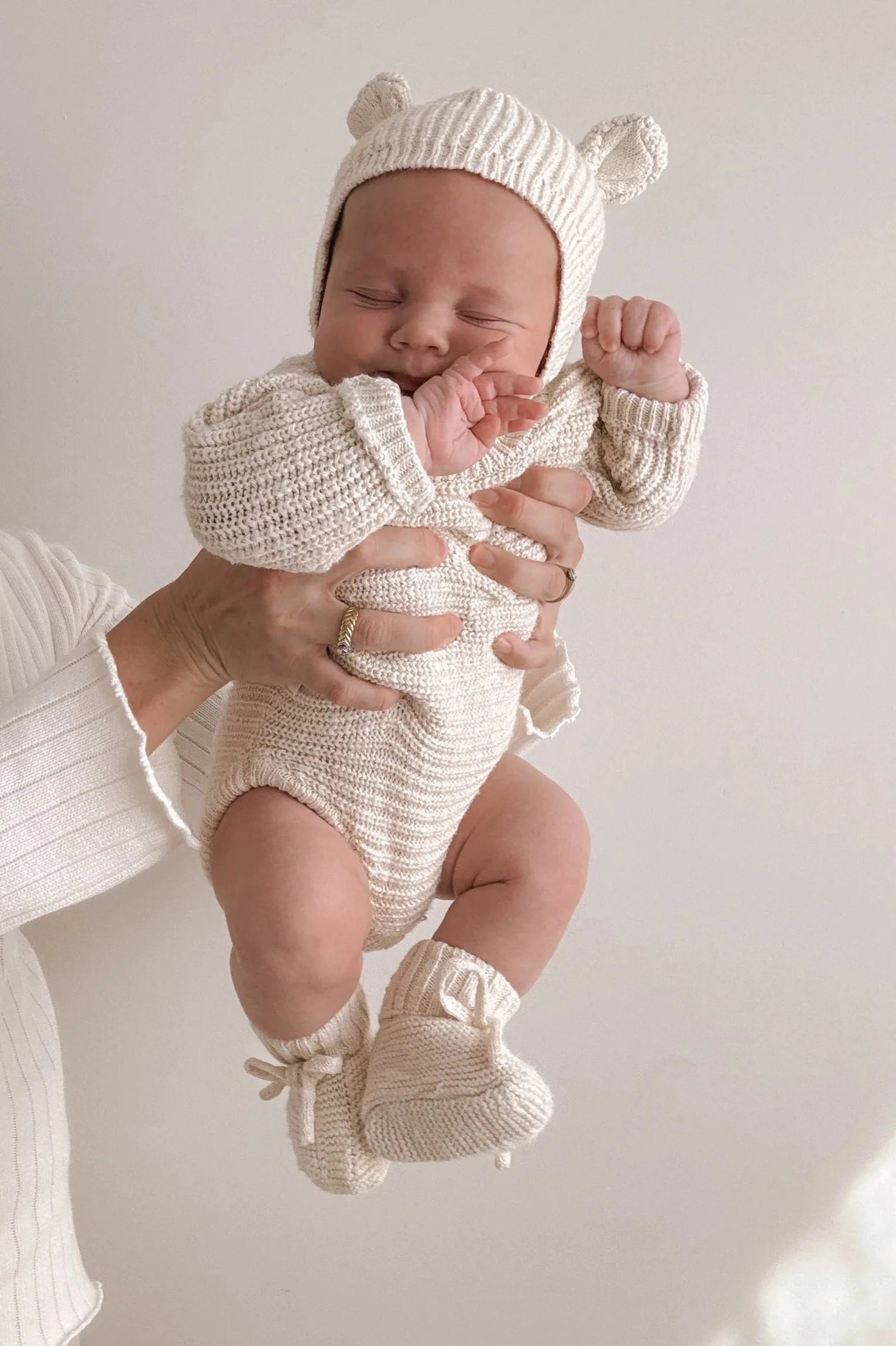 An adult holds a baby against a light-colored background. The baby is dressed in a knitted cream-colored outfit, complete with a hat adorned with small ears, a long-sleeve onesie, and Booties Honey from ZIGGY LOU made of 100% textured cotton. With eyes closed and one hand near their face, the baby looks like an heirloom piece come to life.