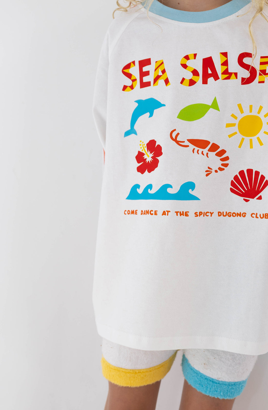 A close-up of a child wearing the SPICY DUGONG Sea Salsa Long Sleeve Tee made from 100% cotton, featuring vibrant illustrations of a dolphin, leaf, sun, wave, shrimp, and shell. The tee displays the text "SEA SALSA" and "COME DANCE AT THE SPICY DUGONG CLUB." The child's lower face and head remain out of view.