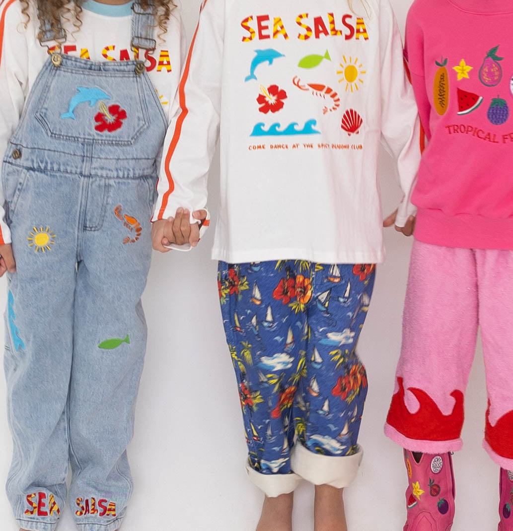 Three children holding hands, all dressed in colorful outfits. The child on the left wears denim overalls and a white long-sleeve tee with "SEA SALSA" and marine-themed prints from SPICY DUGONG. The middle child sports blue pants with red floral prints and a matching shirt adorned with "Fun Sea Salsa print". The child on the right wears a pink long-sleeve shirt with "TROPICAL FRUITS".