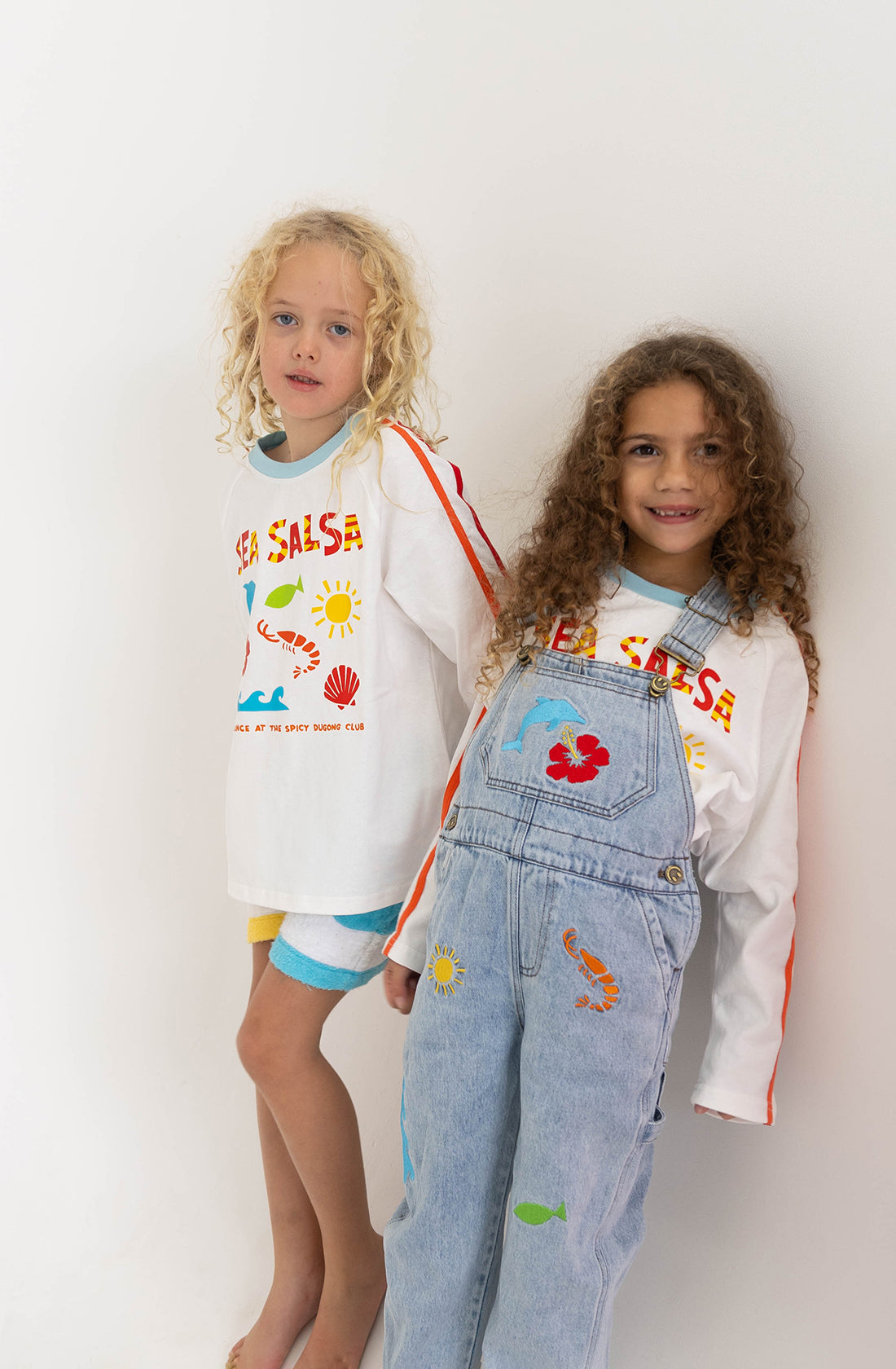 Two children are standing against a plain white background. Both are wearing SPICY DUGONG's Sea Salsa Long Sleeve Tees, made from 100% cotton and featuring colorful Fun Sea Salsa prints. The child on the left has blonde curly hair and is wearing shorts, while the child on the right has brown curly hair and is wearing denim overalls.