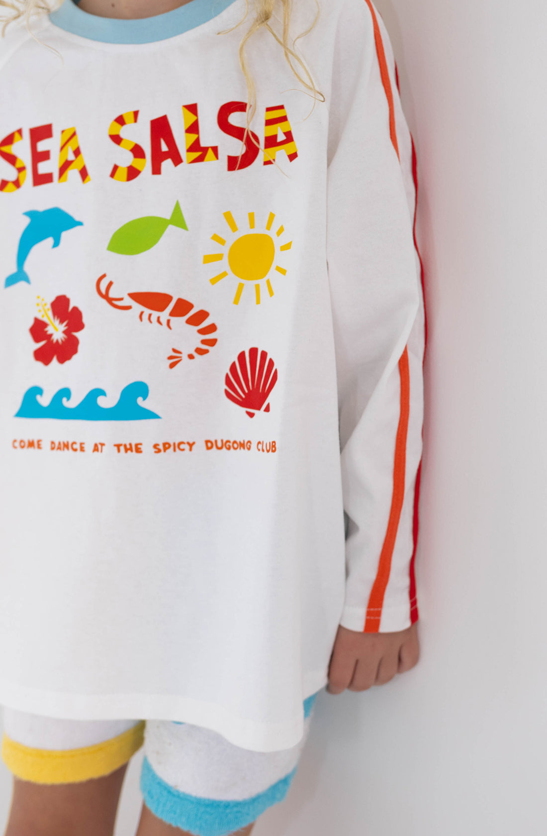 A child wearing the Sea Salsa Long Sleeve Tee from SPICY DUGONG, featuring an exciting print of marine animals, the sun, and objects along with red and orange striped sleeves. Made from 100% cotton, the shirt showcases vibrant graphics and includes text that reads "SEA SALSA" and "COME DANCE AT THE SPICY DUGONG CLUB." The child's face is not visible.