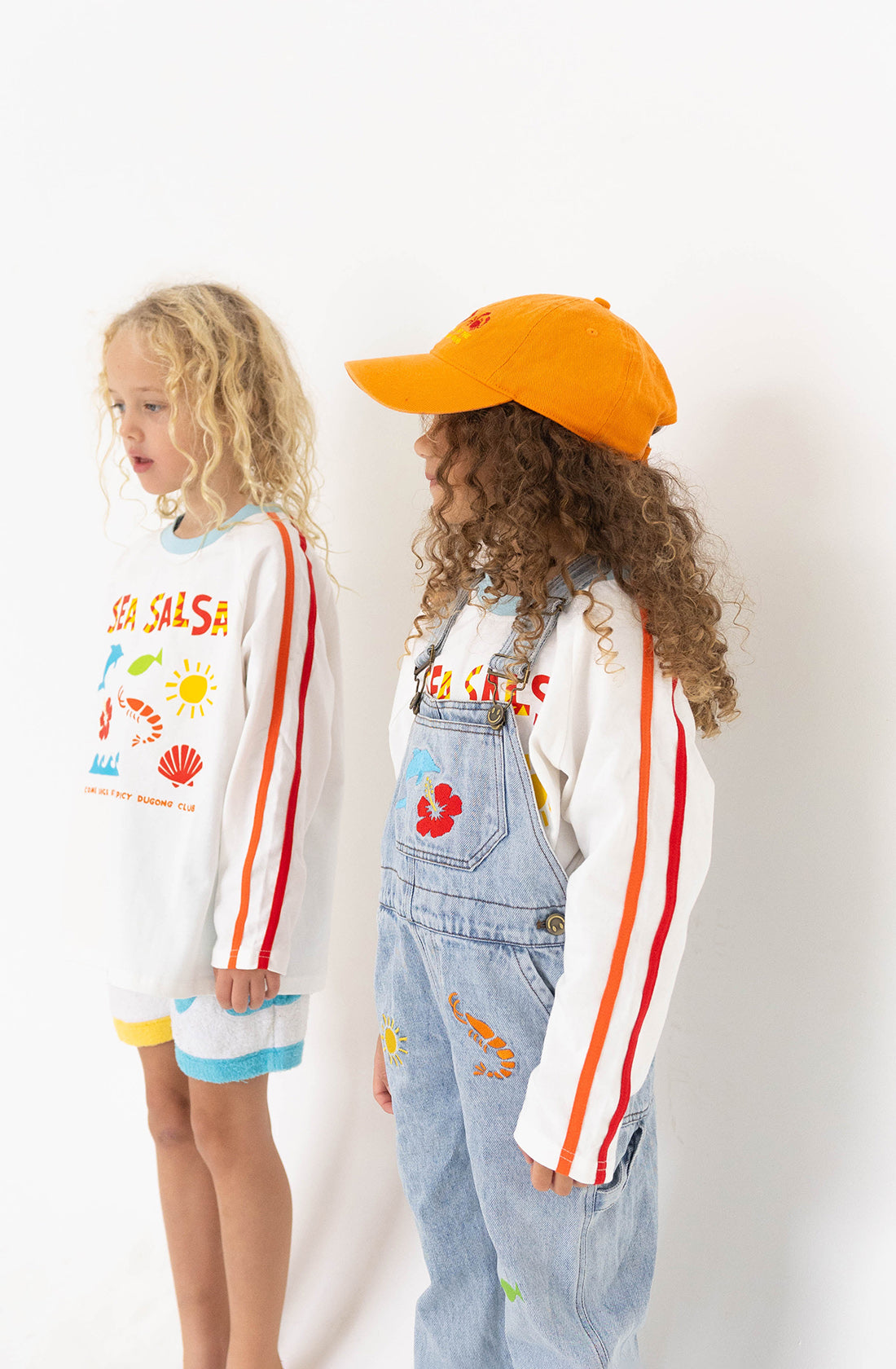 Two children stand against a plain background. The child on the left is wearing a 100% cotton, long sleeve tee with colorful graphics and striped shorts. The child on the right sports SPICY DUGONG's Sea Salsa Long Sleeve Tee featuring a Fun Sea Salsa print, paired with overalls and an orange cap. Both have curly hair.