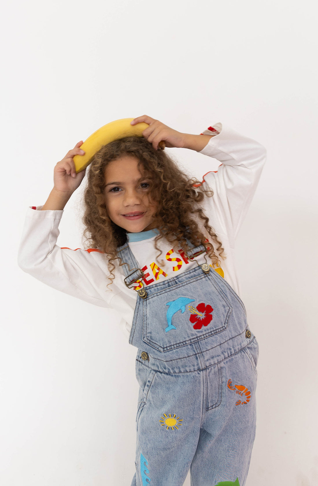 A child with curly hair is smiling while holding a banana over their head. They are wearing the SPICY DUGONG Sea Salsa Long Sleeve Tee made of 100% cotton and denim overalls decorated with colorful patches, including flowers and animals. The fun sea salsa print adds an extra touch of whimsy to the ensemble against the plain white background.