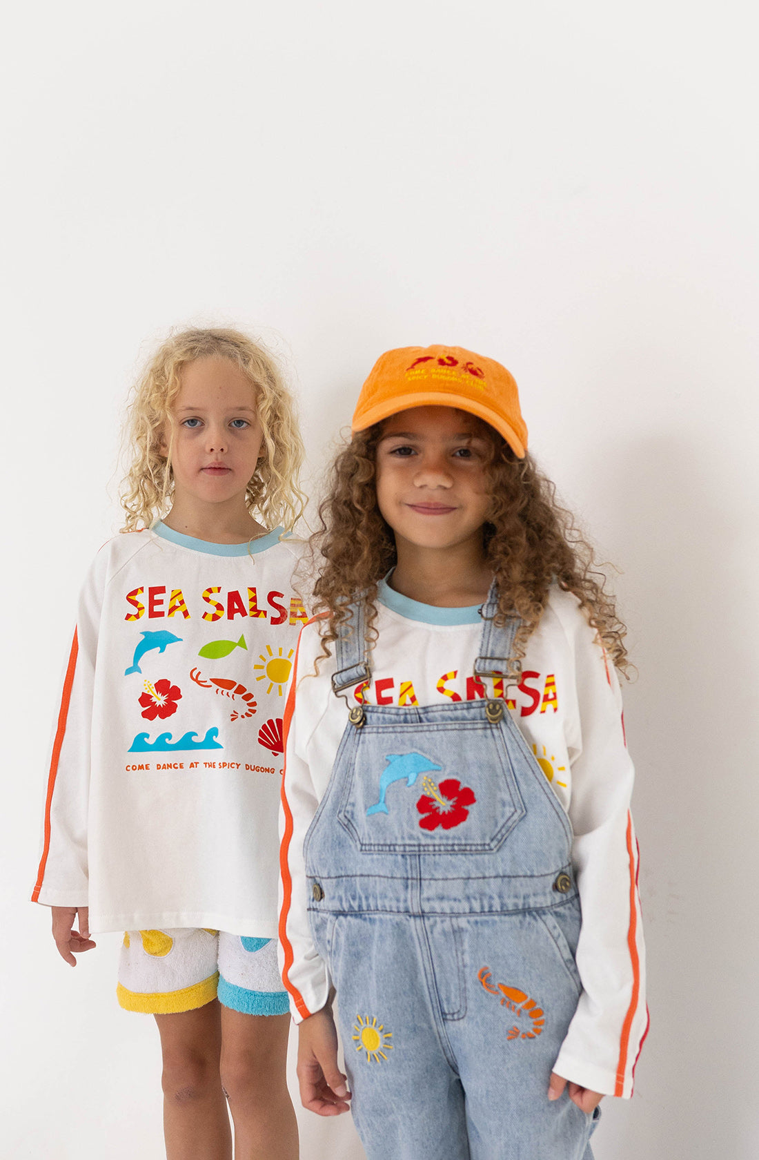 Two children standing in front of a white background. The child on the left, wearing the SPICY DUGONG Sea Salsa Long Sleeve Tee made of 100% cotton featuring colorful text and images, paired with yellow-blue shorts, has curly blonde hair. The child on the right, sporting a white shirt under denim overalls and an orange hat, showcases the Fun Sea Salsa print with marine-themed graphics.