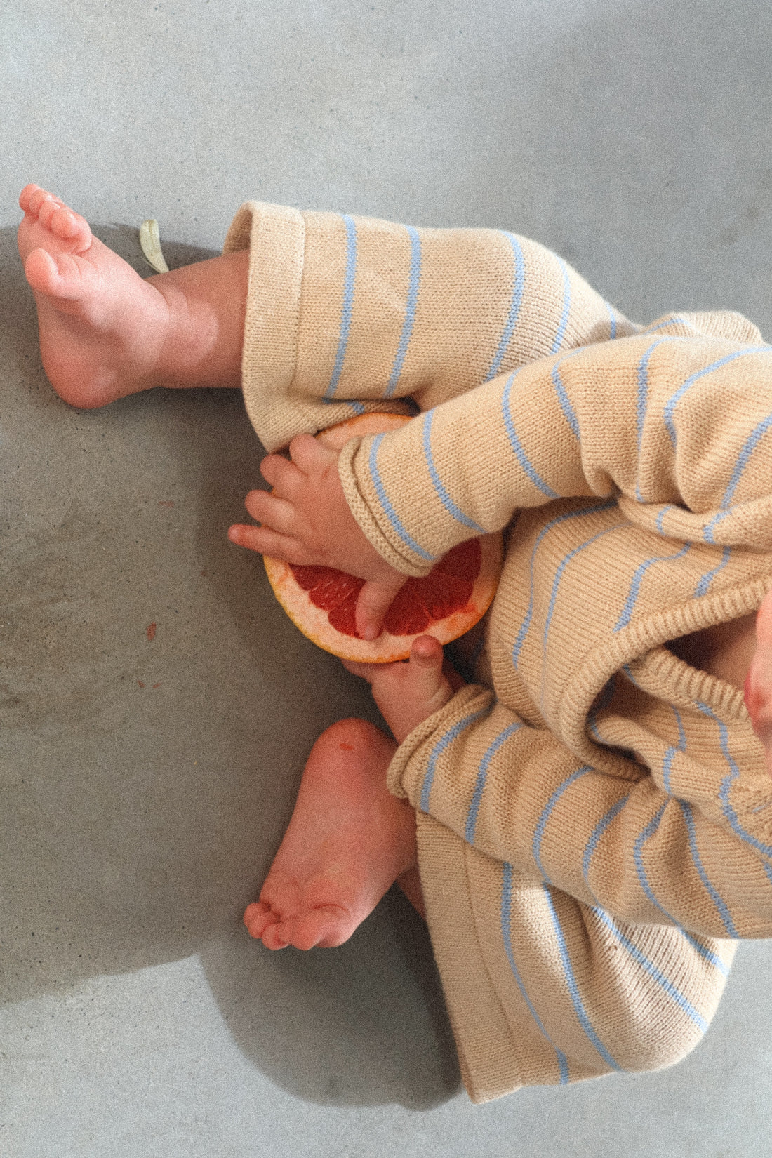 A baby, dressed in MILKY DESIGNS' Staple Pant Blue Stripe outfit made of 100% cotton knit, sits on a grey surface holding a halved grapefruit. The baby’s legs and feet are visible with one leg placed over the other, showcasing the outfit's comfortable 3/4 fit and elastic waistband near the baby's lap.