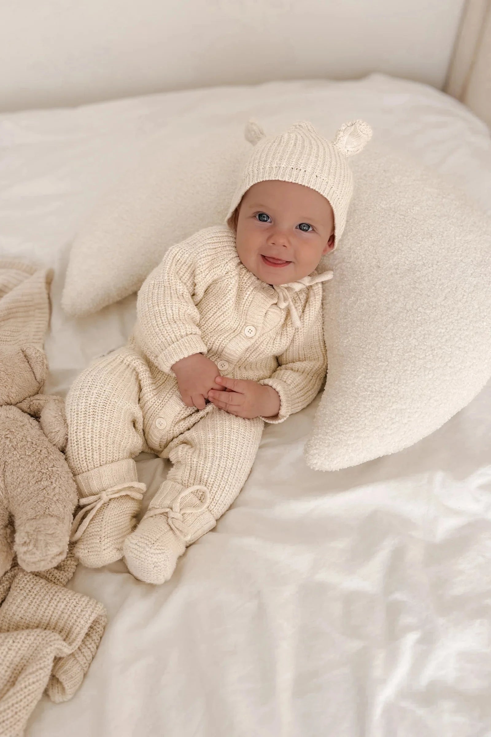 A smiling baby wearing a cream-colored knitted outfit with matching hat featuring tiny bear ears and ZIGGY LOU's Booties Honey lies on a cream-colored pillow and bed. The baby has a plush blanket nearby and rests comfortably, making the scene an heirloom piece to cherish.