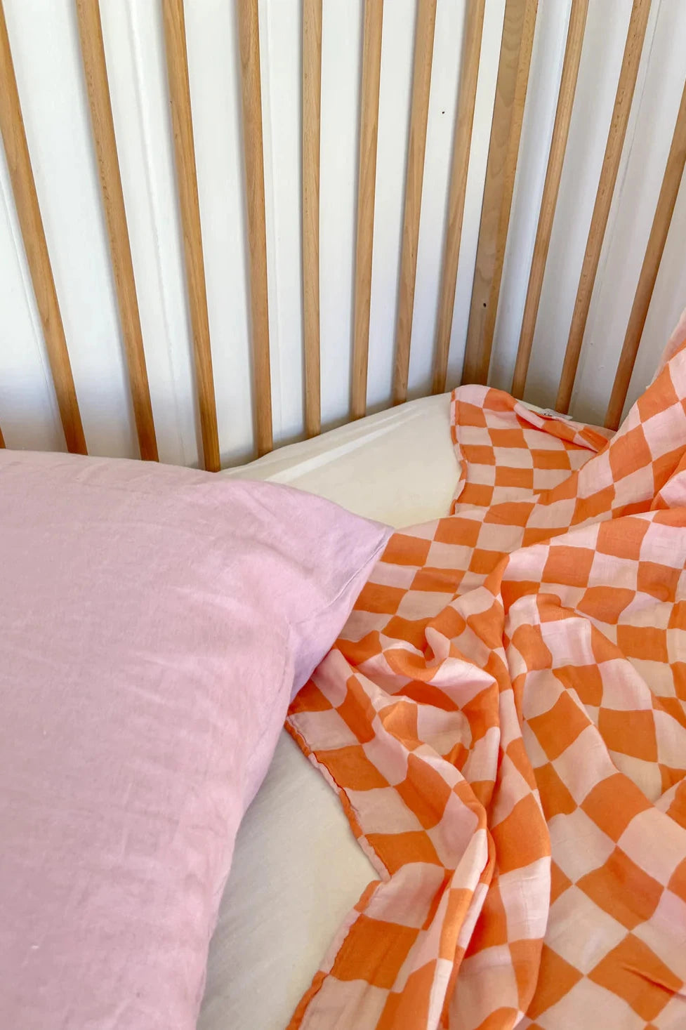 An image of a simple wooden crib with vertical slats. Inside, a pink pillow on a white mattress rests under an orange and white checkered Apricot Peaches Wrap, partially spread out and slightly wrinkled, showcasing MILKY DESIGNS' unique textile prints. The background features plain white walls.