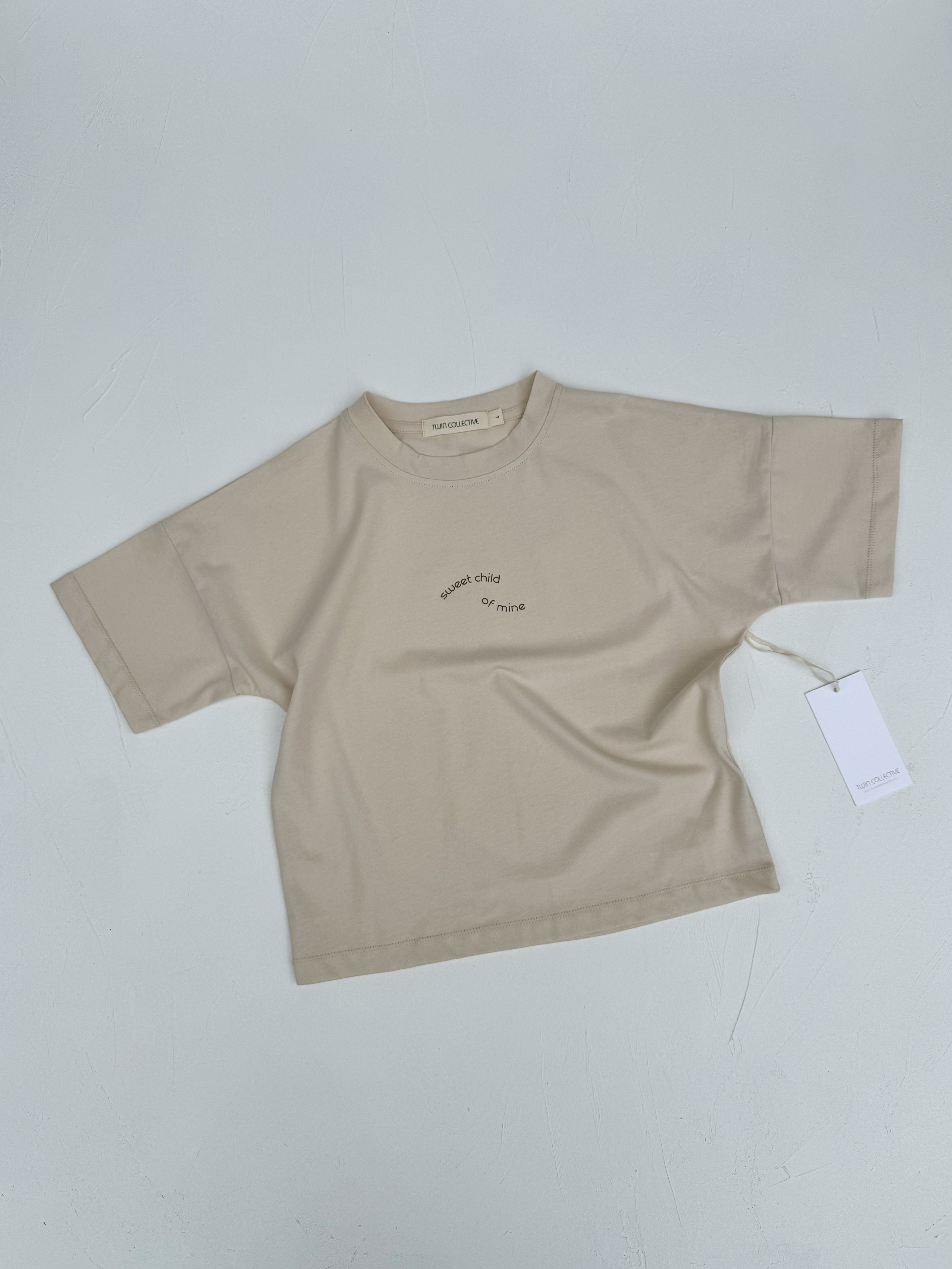 The Sweet Child Tee from TWIN COLLECTIVE is a beige, oversized kids' short-sleeve shirt made of 100% cotton and designed with an unfinished sleeve style. It features the words "WINNER CHILD of WINNER" printed in an arc on the chest. The tee is displayed flat on a light gray surface with a white tag attached to the right sleeve.