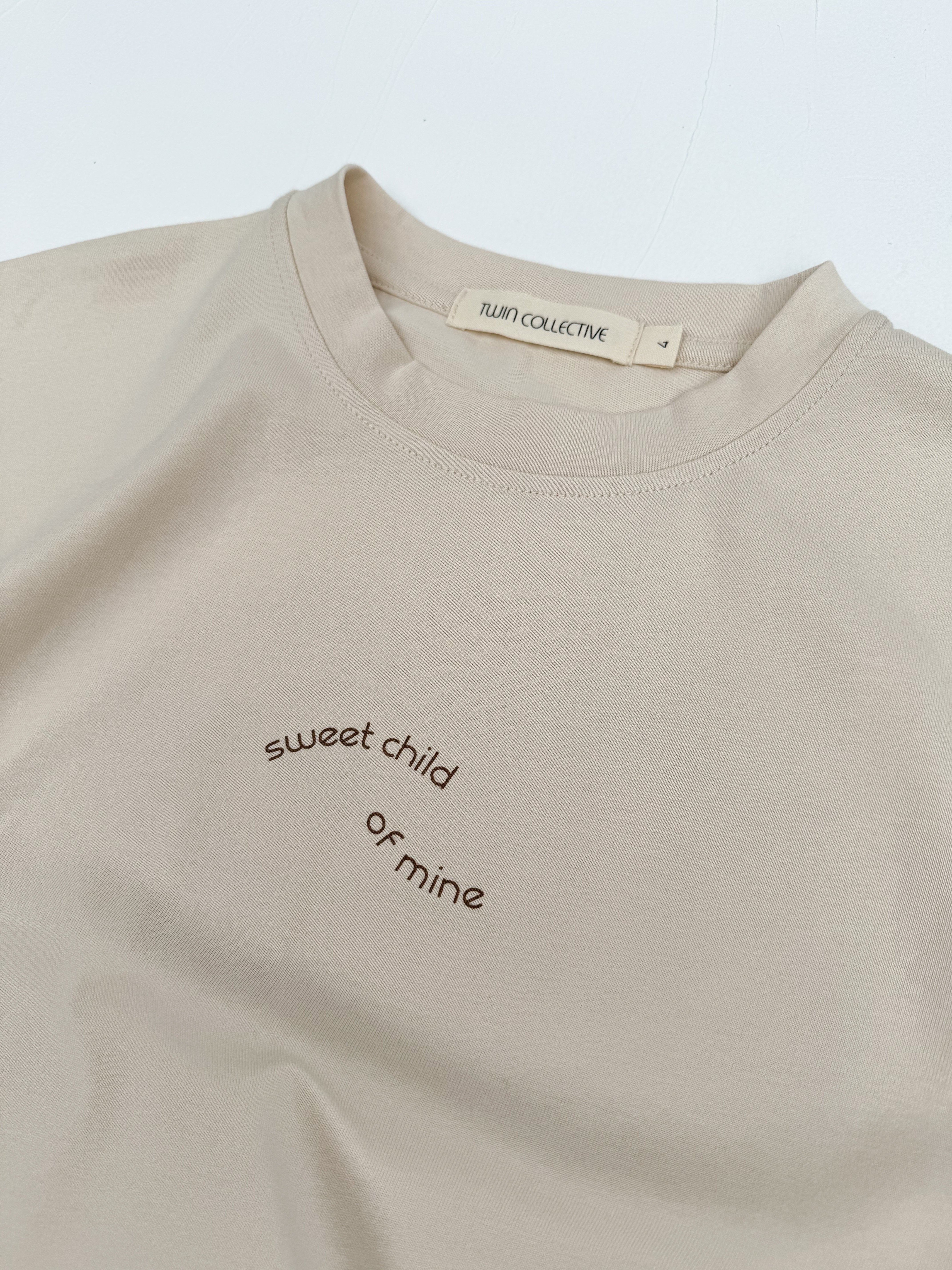 A light beige T-shirt, called the Sweet Child Tee, is laid flat on a white surface. The kids’ tee, made from 100% cotton, features minimalist text in a small, curved font that reads "sweet child of mine" across the chest. Designed with an oversized fit for extra comfort, a tag inside the collar displays the brand name "TWIN COLLECTIVE.