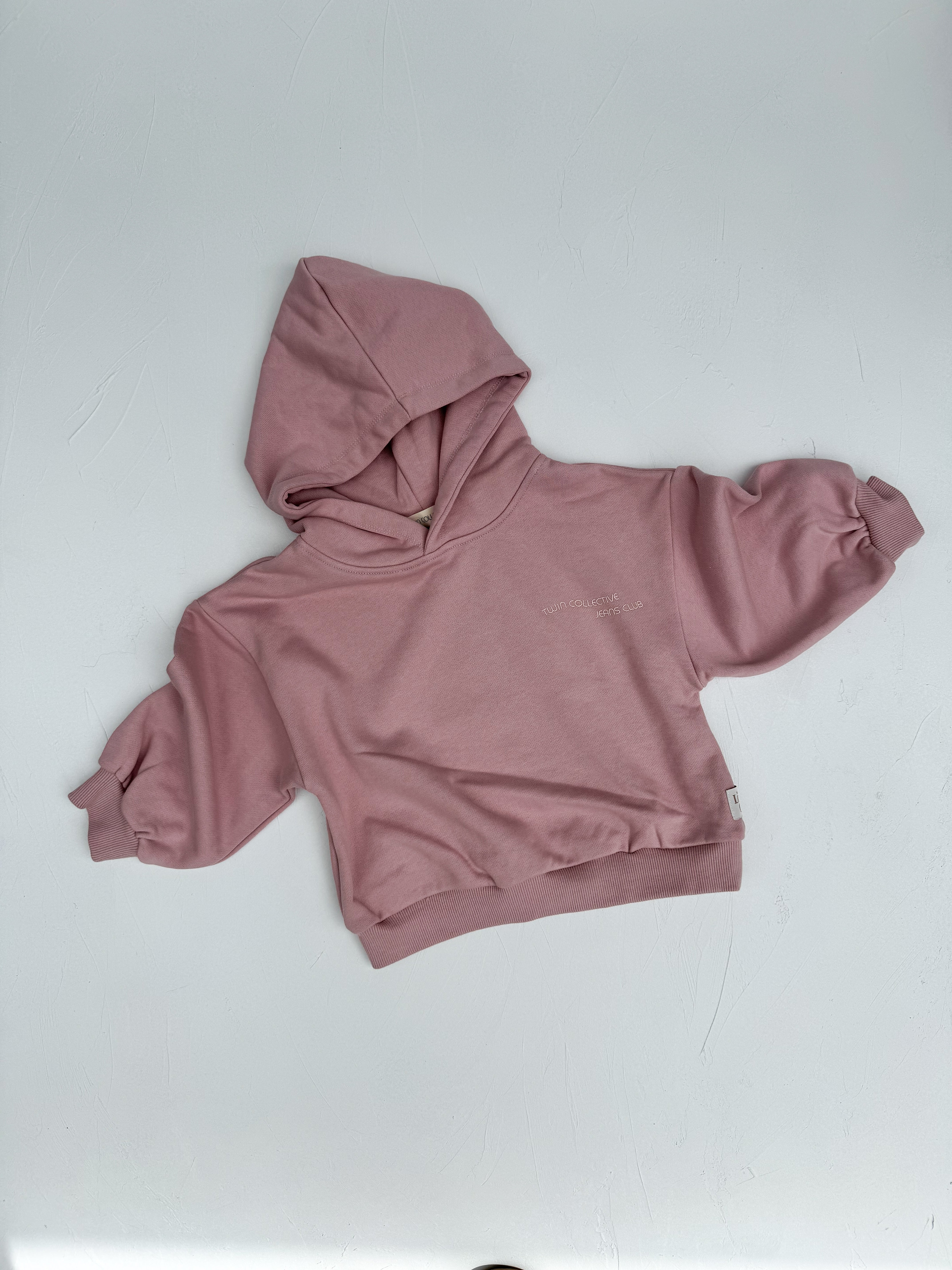 The Logo Hoodie Set Pink Haze by TWIN COLLECTIVE, featuring a small pink hooded sweatshirt designed with heavy loopback terry fleece, is laid flat on a white surface. This cozy garment boasts long sleeves, a front pocket, and a hood, perfectly embodying the essence of comfort and style. Ideal for a child.