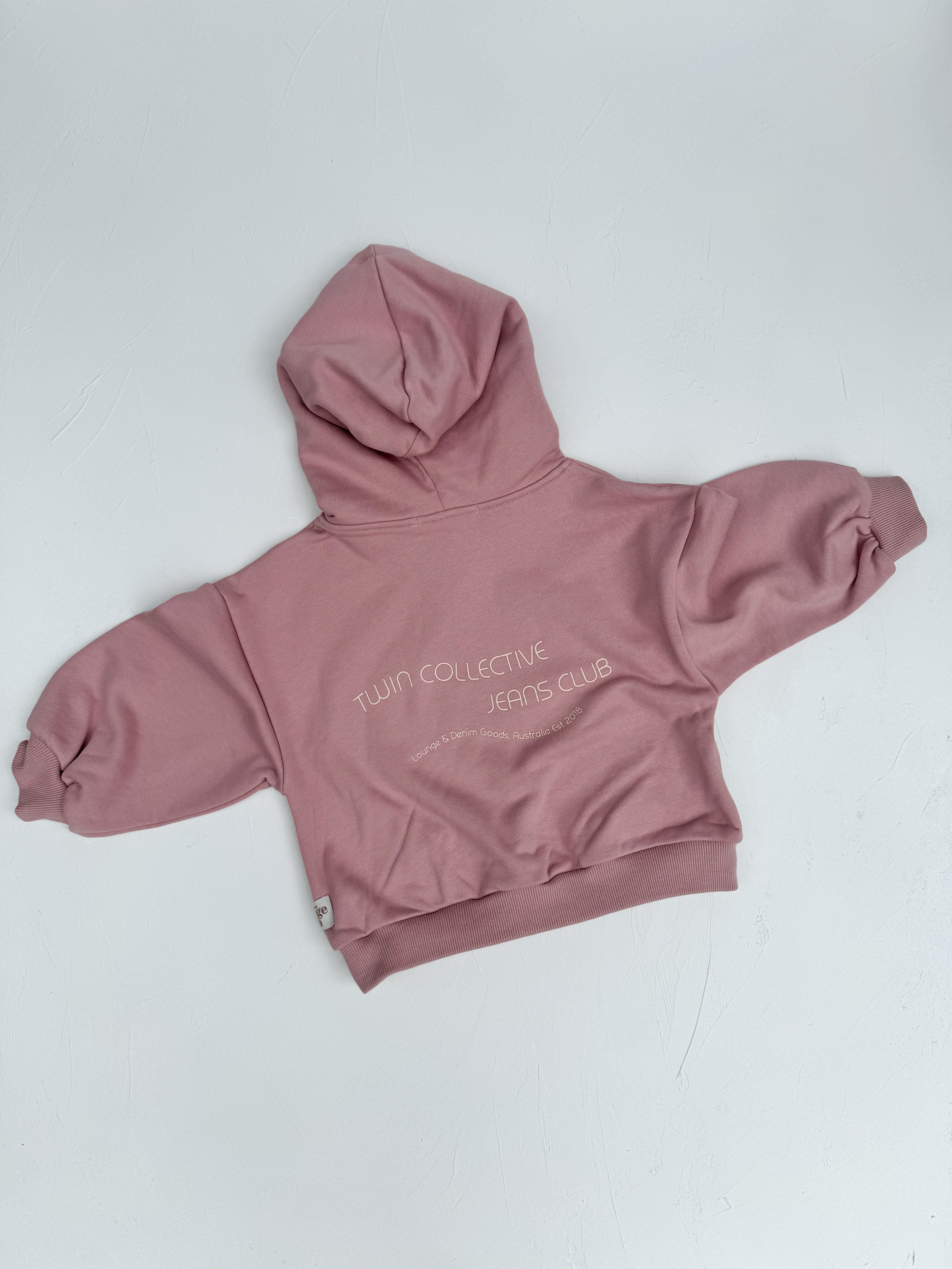 A "Logo Hoodie Set Pink Haze" from TWIN COLLECTIVE lies flat with its back side facing up. The text "TWIN COLLECTIVE JEANS CLUB" is printed in white on the back. Made from heavy loopback terry fleece, the sweatshirt has long sleeves with cuffs, a waistband, and a small tag on the lower left side.