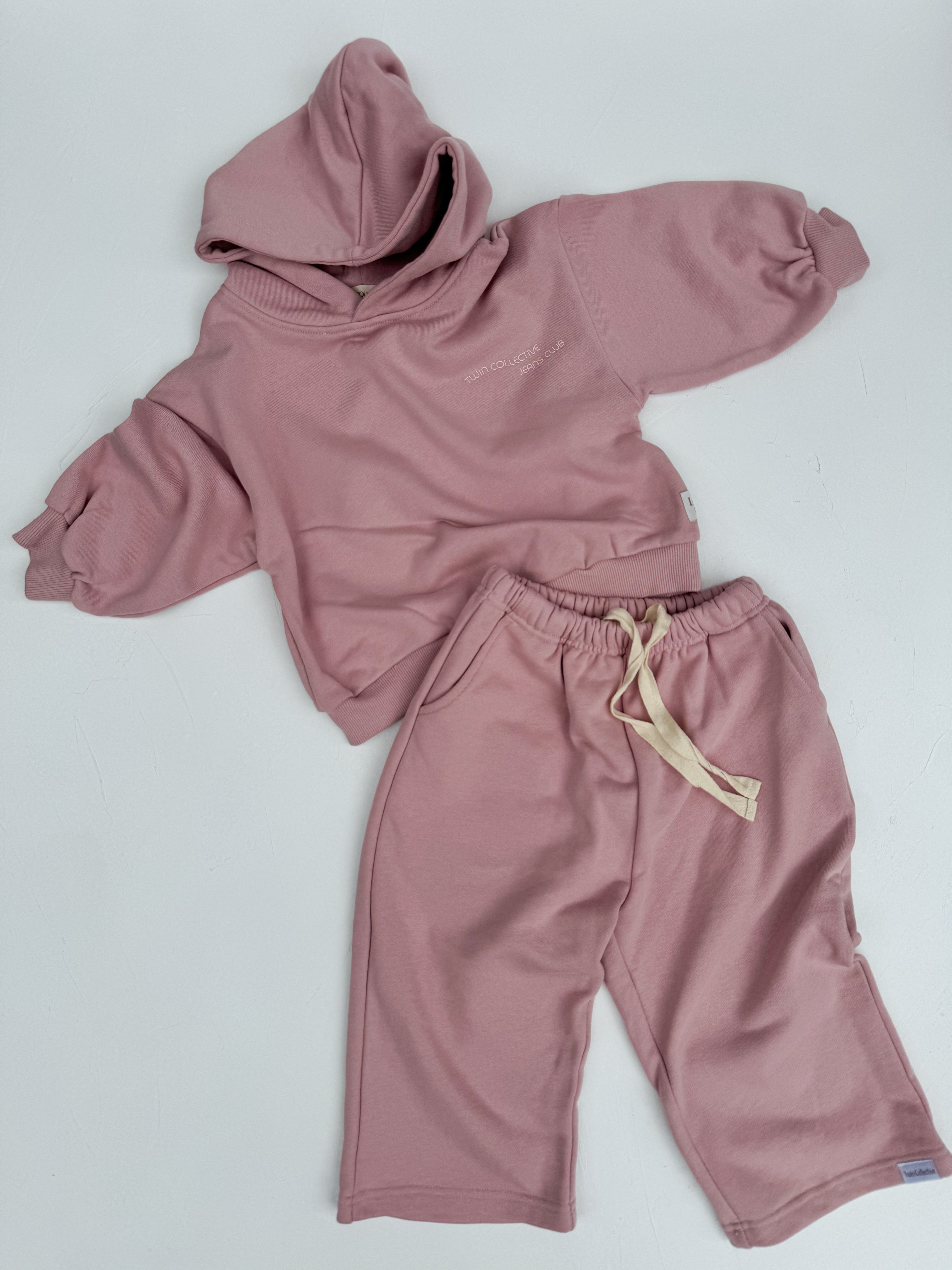 A children's outfit from TWIN COLLECTIVE featuring a light pink hooded sweatshirt with a small embroidered design on the chest paired with Lounge Track Pants in Pink Haze. The oversized track pants include an elastic waistband and a beige drawstring. Both pieces are elegantly laid out on a white surface.