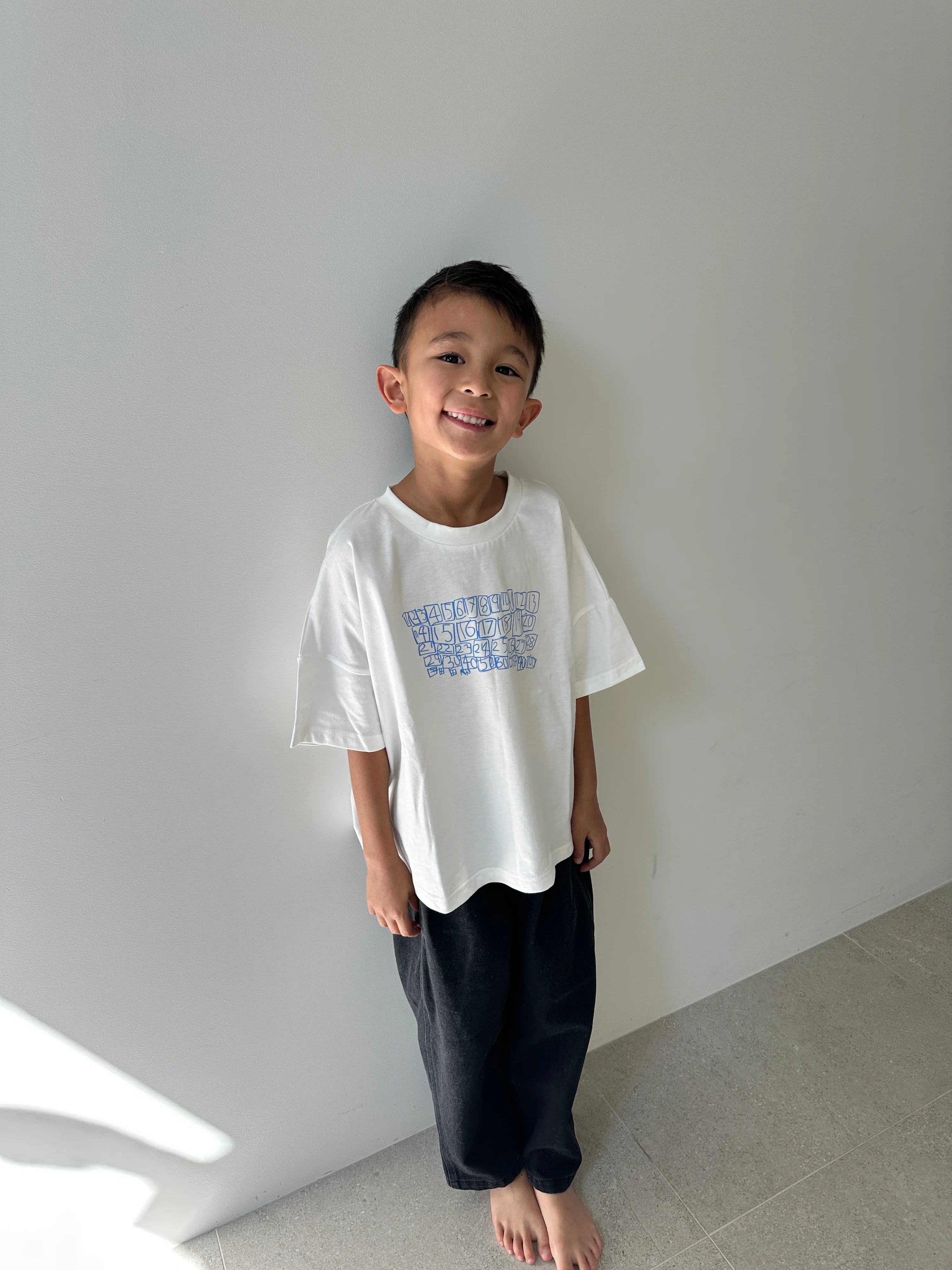 A young boy with short dark hair stands barefoot indoors against a light wall, smiling and wearing the TINY TROVE Parker Numbers Relaxed Tee White/Blue and loose black pants. Sunlight falls on the floor beside him.
