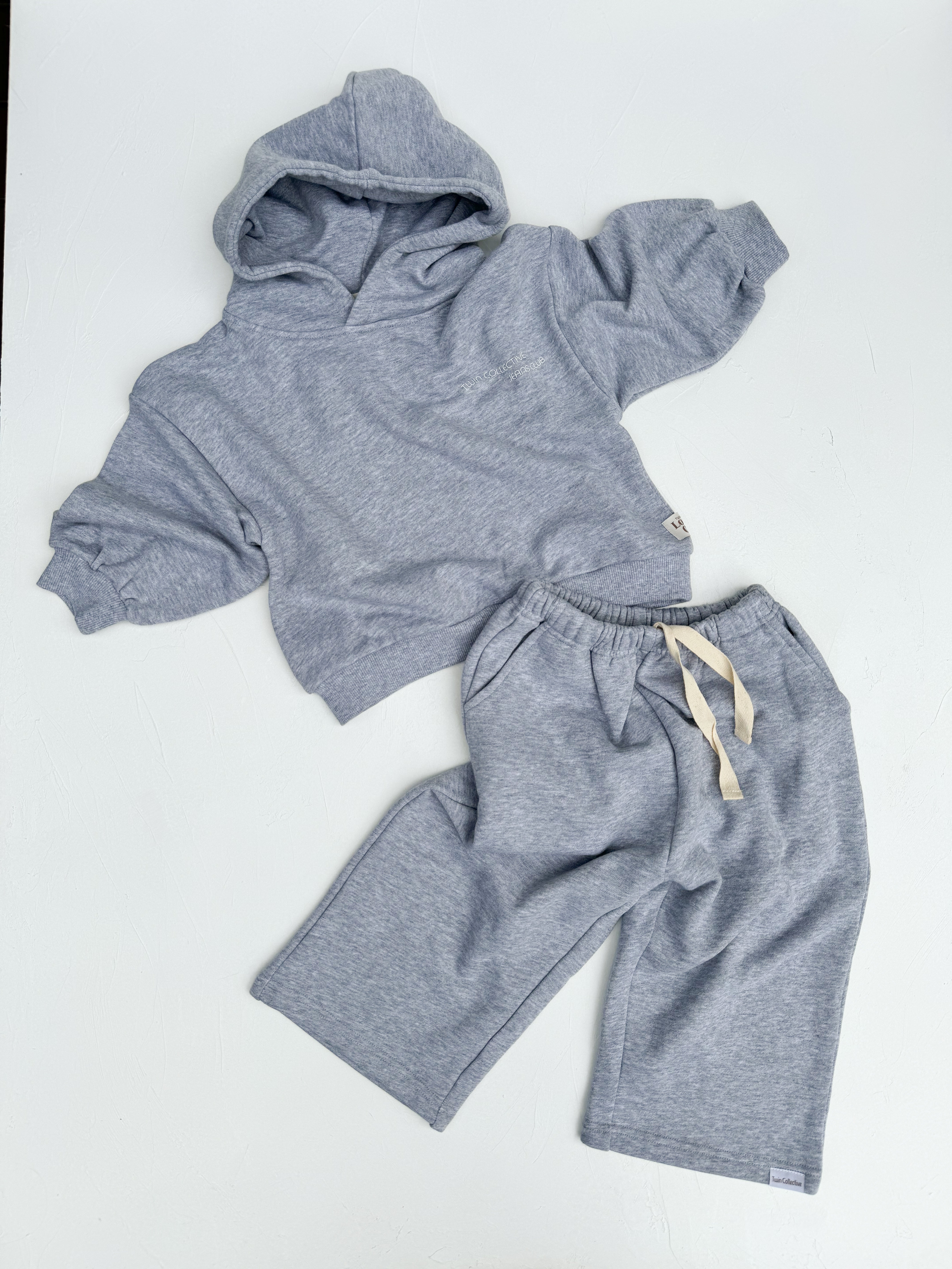 A cozy, gray children's outfit from TWIN COLLECTIVE is displayed on a white surface. The ensemble includes the Logo Hoodie Set Grey Marle, featuring a Lounge Club Hoodie with long sleeves and a pair of matching elastic-waist pants with a beige drawstring. Crafted from soft Loopback Terry fleece, this set is perfect for casual wear and ultimate comfort.