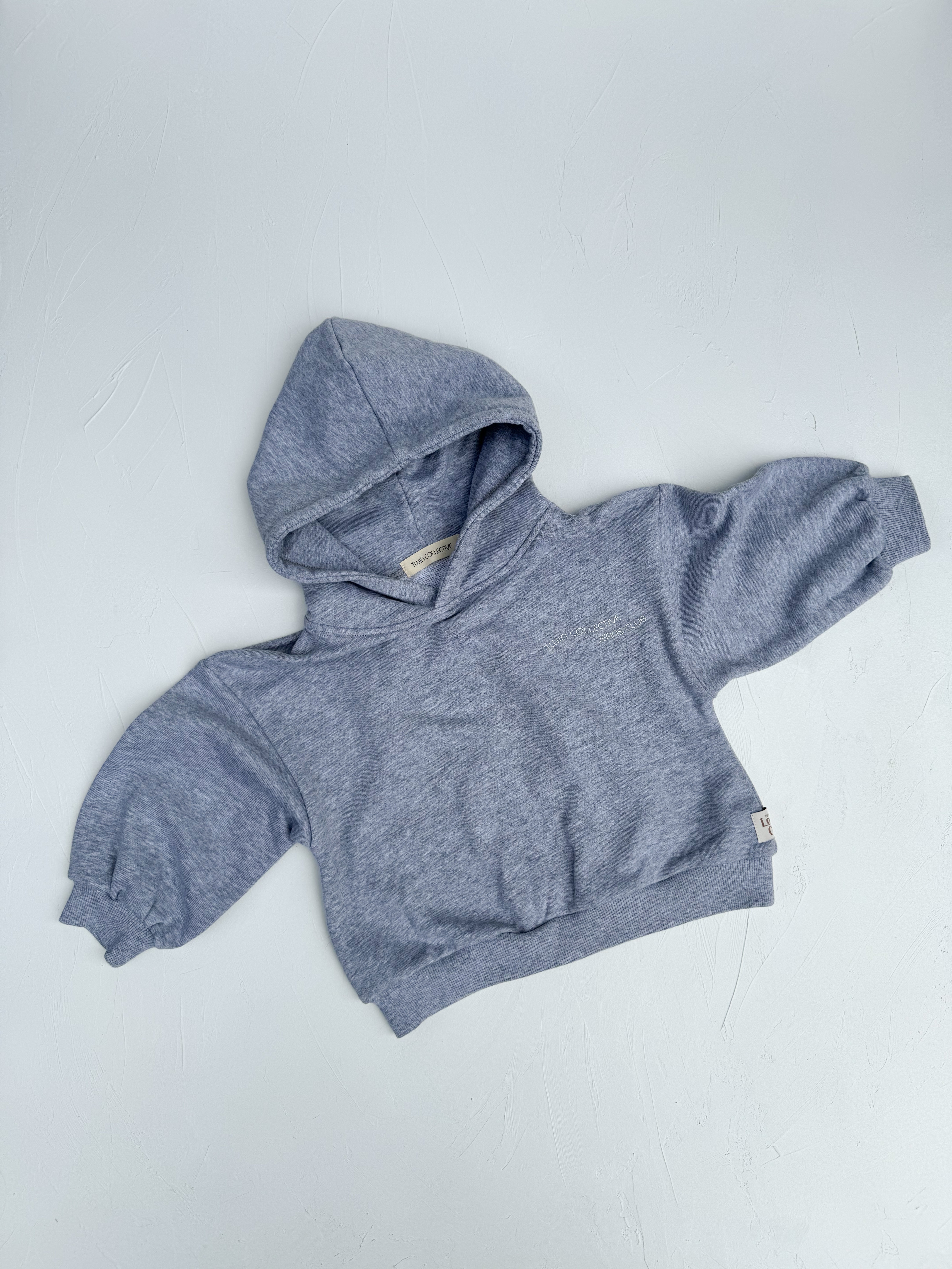 A TWIN COLLECTIVE Logo Hoodie Set in Grey Marle is laid flat against a white background. The light gray hoodie, which features long sleeves and a front pocket, is made of loopback Terry fleece with a heathered texture. The pointed hood adds to its design, which is tailored for a small child.