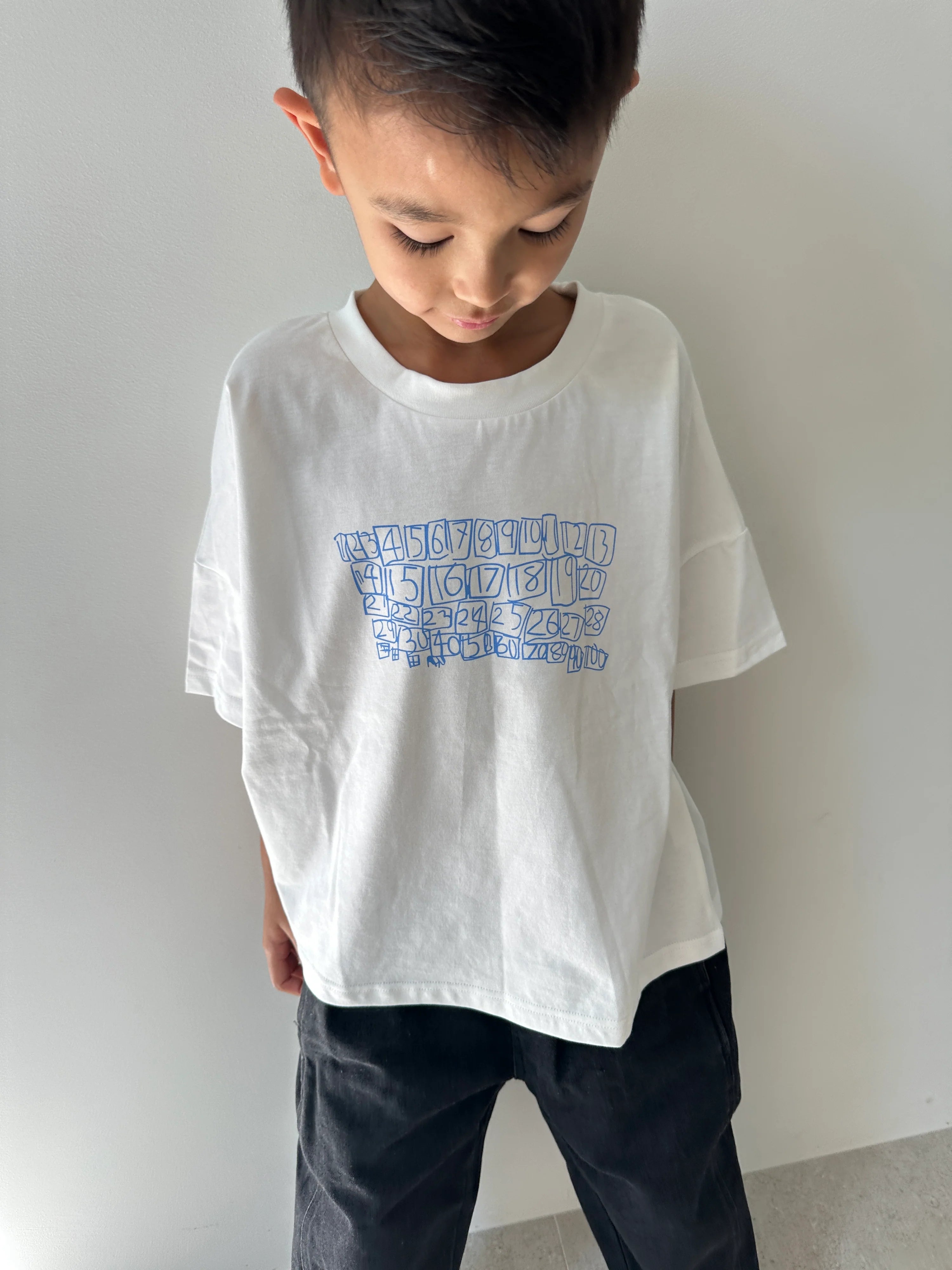 A young boy with short hair looks down, wearing the TINY TROVE Tiny Trove - Parker Numbers Relaxed Tee White/Blue and black pants, standing against a light gray wall.