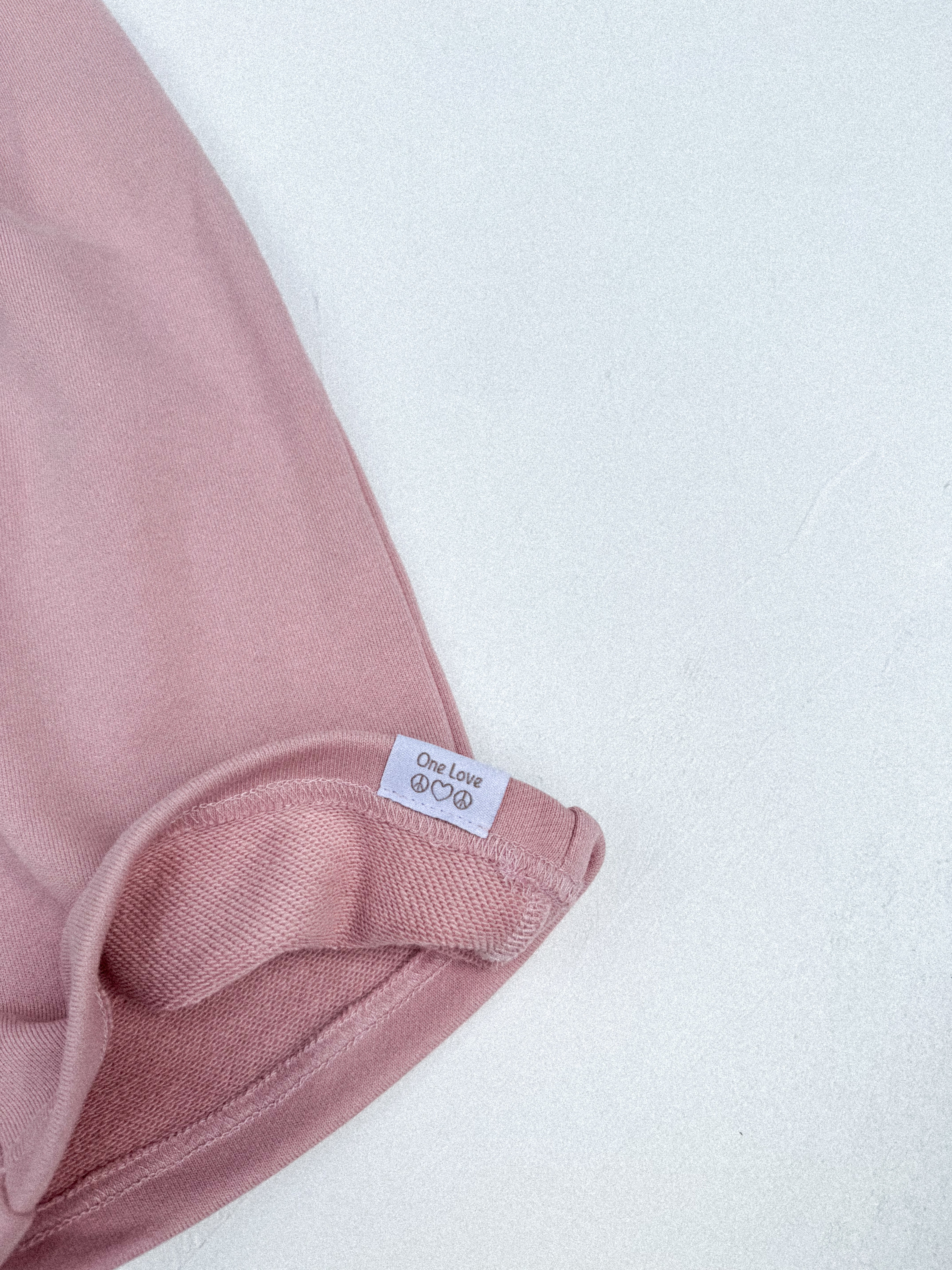 Close-up of the hem of TWIN COLLECTIVE's Logo Hoodie Set in Pink Haze, featuring a small white tag with "One Love" and an infinity symbol. The fabric at the hem appears slightly folded, showcasing the texture of heavy loopback terry fleece against a light background.