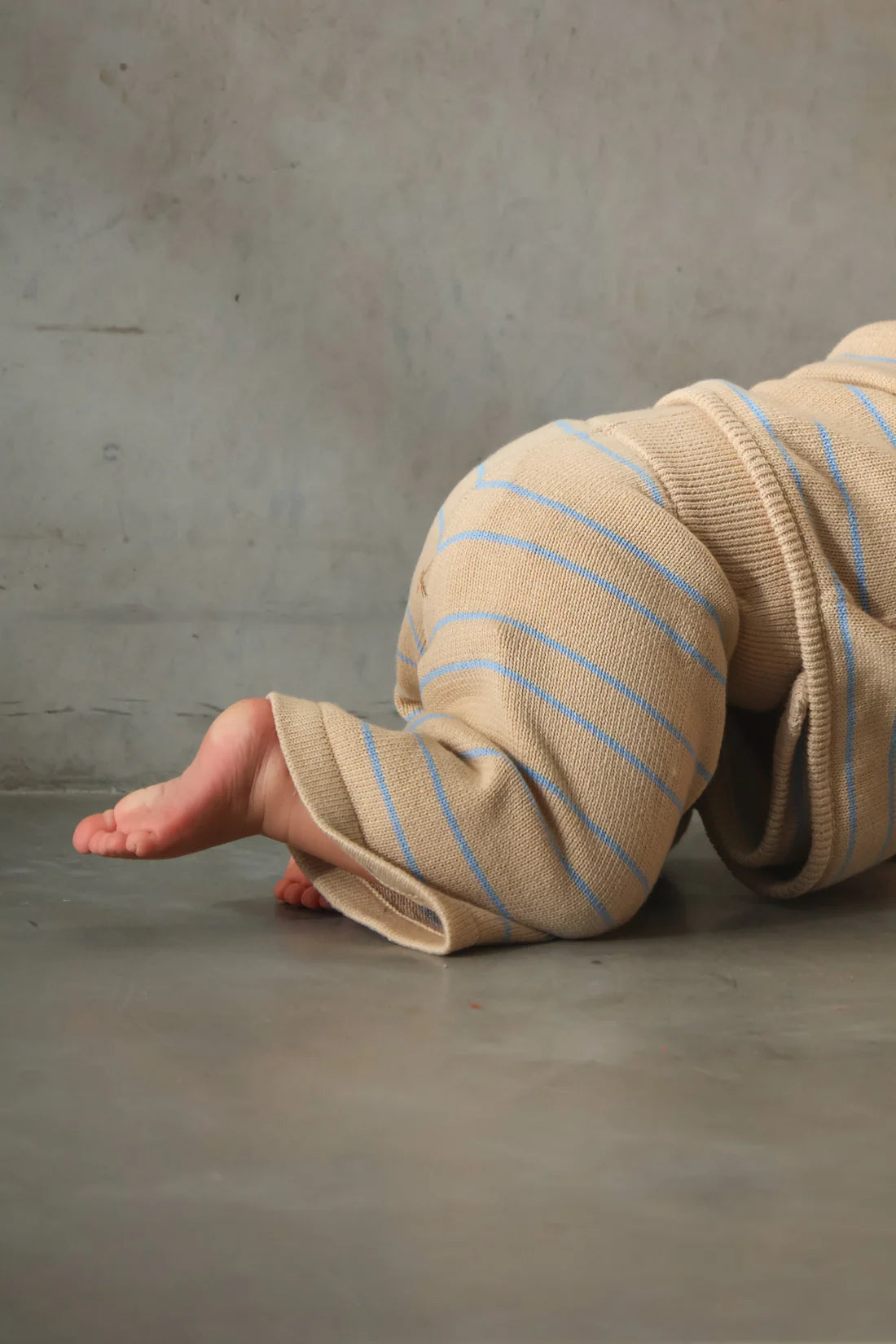 A baby dressed in MILKY DESIGNS' Staple Pant Blue Stripe, featuring beige pants with blue stripes and an elastic waistband, is seen crawling on a smooth, grey floor. The baby’s focus is on moving forward, with legs and feet in motion. The background is a soft, neutral-toned wall.