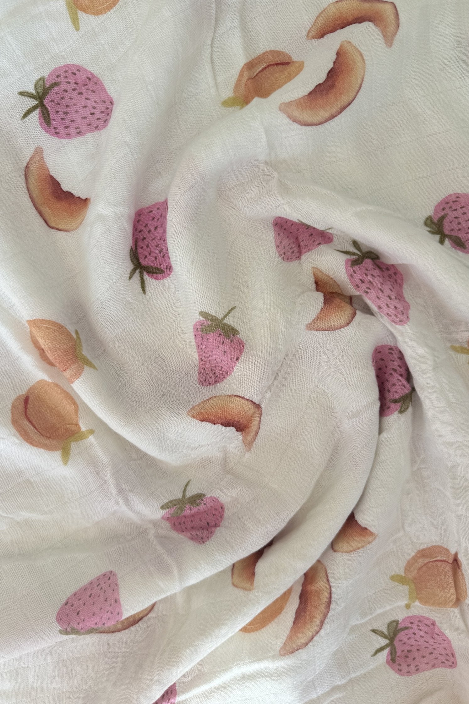 The Fruiti Wrap by MILKY DESIGNS is a white fabric adorned with distinctive textile prints of pink strawberries with green leaves and peach slices in shades of peach and brown. Known for its buttery softness, this slightly wrinkled fabric is perfect for versatile wraps.