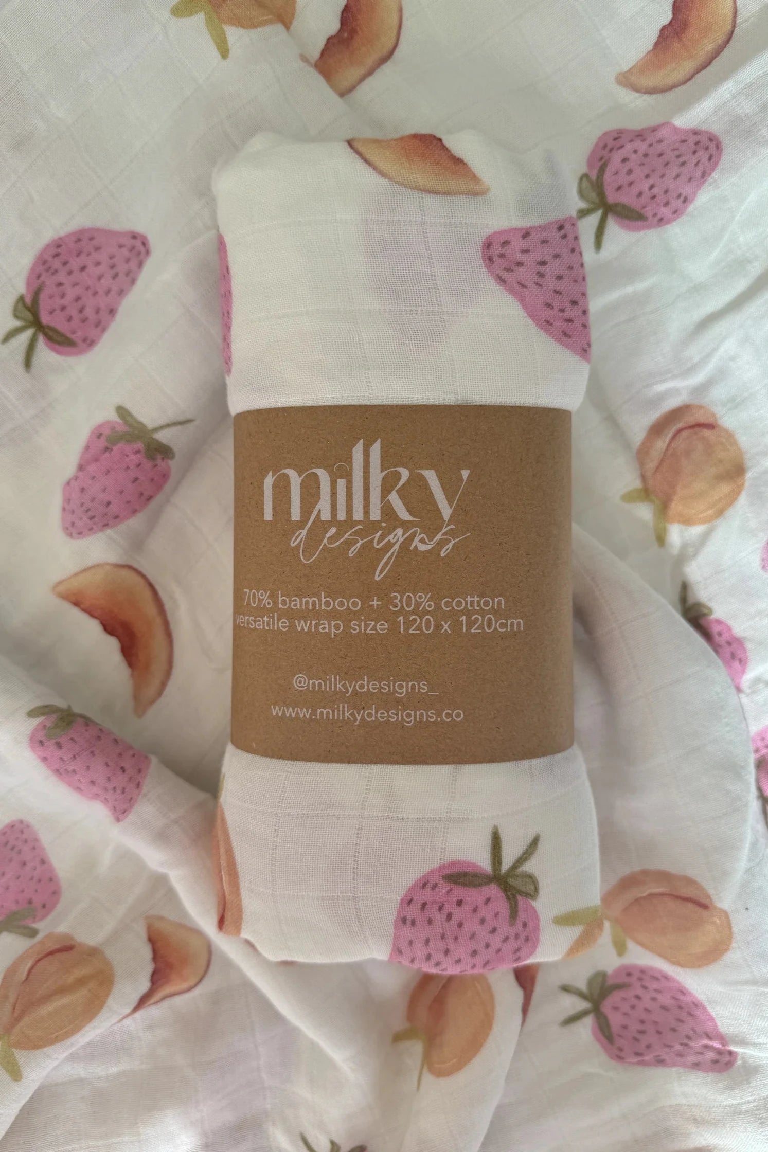 A rolled-up Fruiti Wrap with a label reading "MILKY DESIGNS" rests on top of the unrolled fabric. This versatile wrap is adorned with a pattern of pink strawberries and peach halves. The label indicates it boasts unique textile prints, made of 70% bamboo and 30% cotton, measuring 120x120 cm.