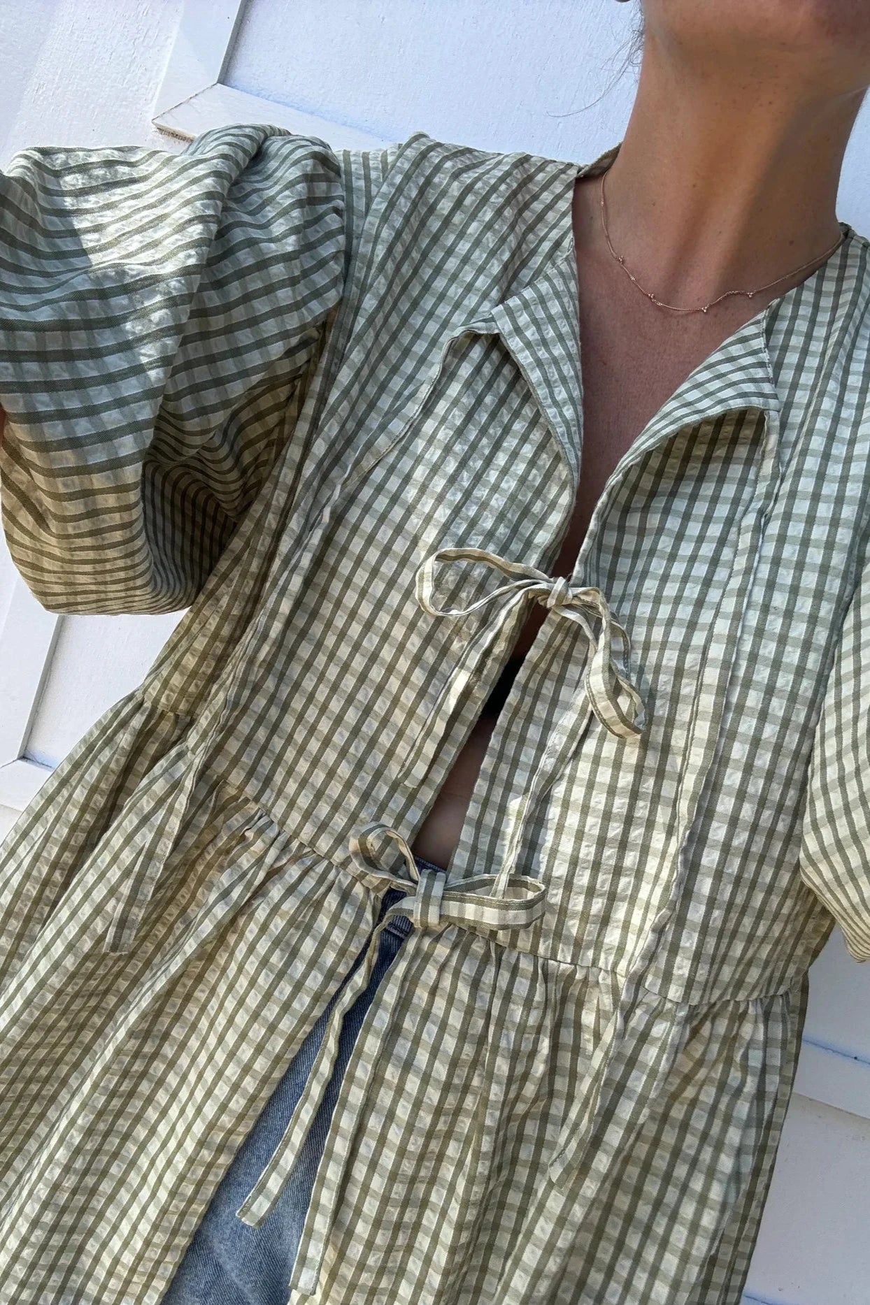 A person wears the Mama Cami Blouse in Olive Gingham by LITTLE THE LABEL, featuring puffed sleeves and front ties. The relaxed fit beautifully complements the green and white checkered pattern. The white wall close-up background enhances the cozy yet stylish appeal.