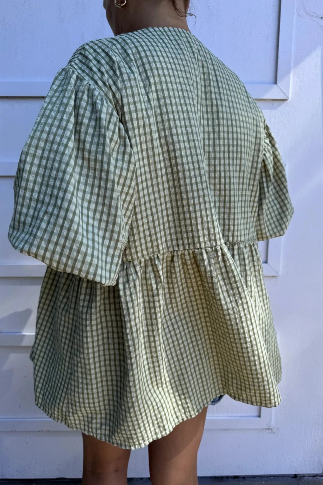 A person wearing the Mama Cami Blouse in olive gingham from LITTLE THE LABEL stands in front of a white wall. The blouse, featuring a loose-fitting design and puffy sleeves, complements the person's tied-back hair elegantly.