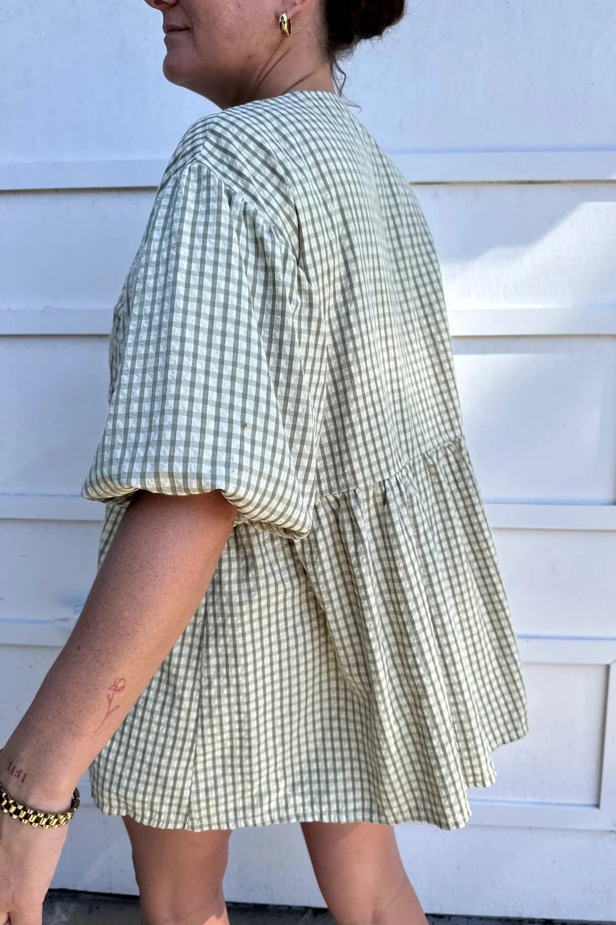 The woman is dressed in a Mama Cami Blouse Olive Gingham from LITTLE THE LABEL, featuring an olive and white gingham pattern with short sleeves. She accessorizes with gold hoop earrings and a bracelet, reveals a small tattoo on her arm, and stands against a white background while slightly smiling.