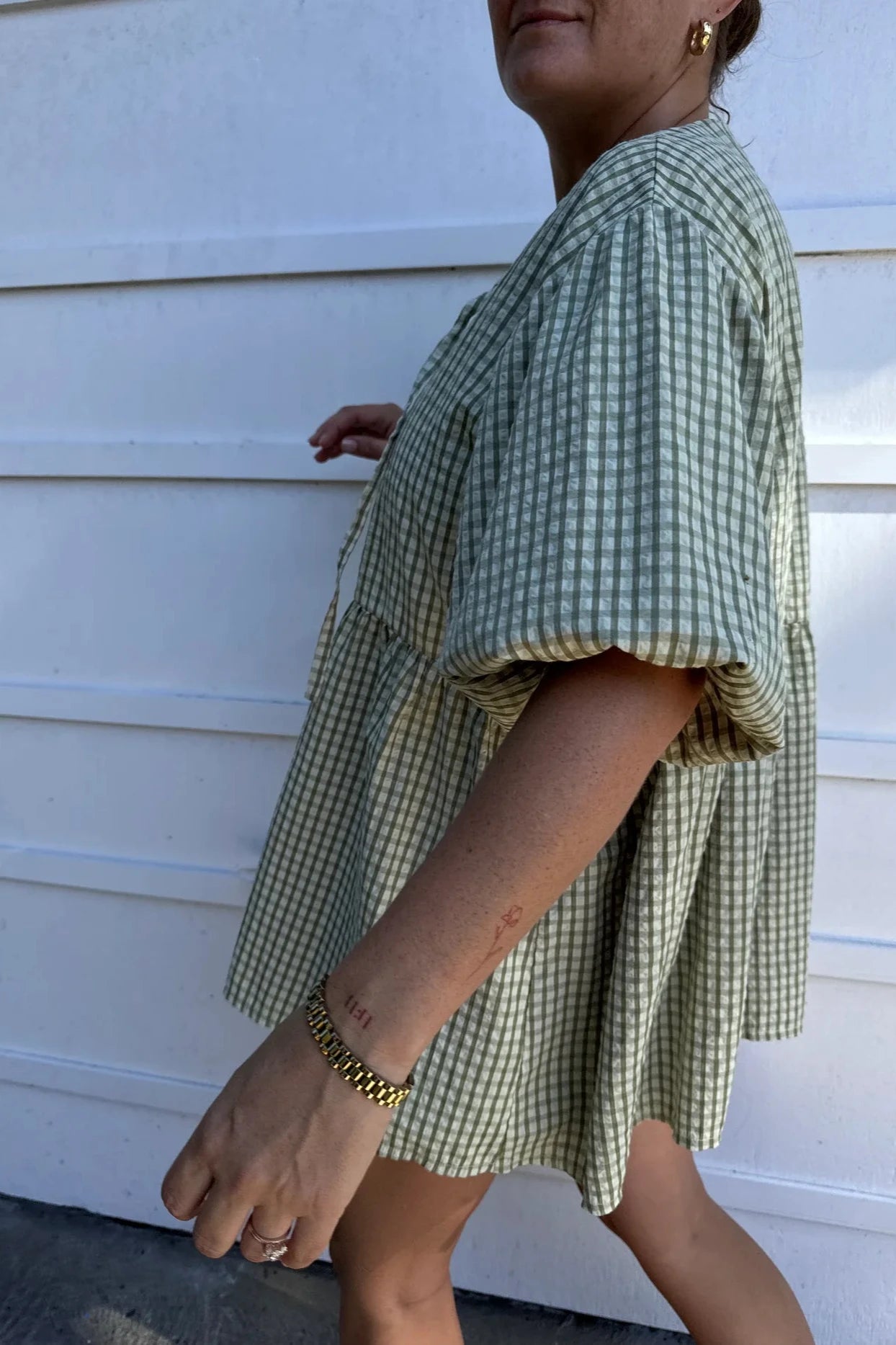 A person wearing the Mama Cami Blouse in olive gingham by LITTLE THE LABEL walks outdoors. The blouse, with its puffed sleeves and A-line design, complements their arm tattoo and bracelet. A white wall serves as the background, highlighting the casual elegance of their outfit.