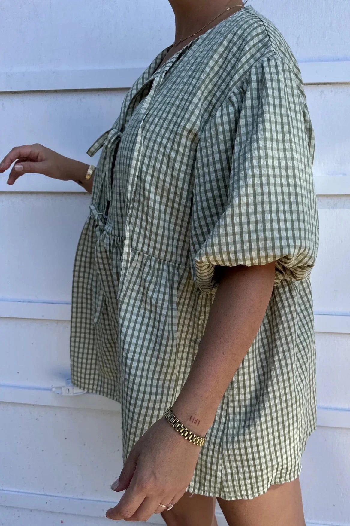 The individual is wearing the Mama Cami Blouse in Olive Gingham, designed by LITTLE THE LABEL, featuring puffed sleeves. They are standing sideways against a white background, showcasing a tattoo on their arm and sporting a gold bracelet. Only the upper body is visible.