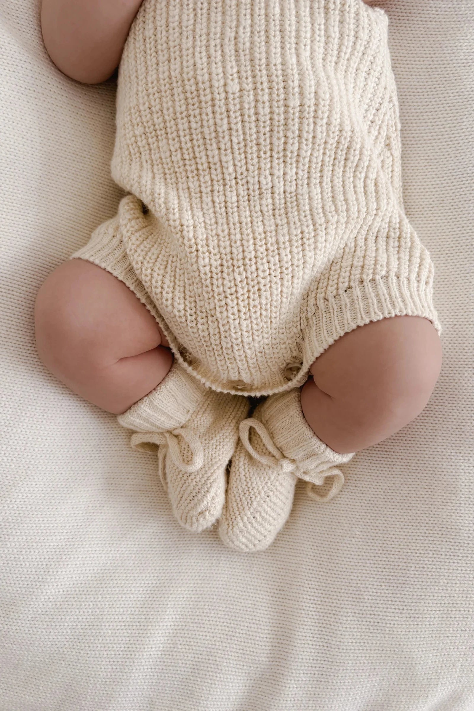 A baby curled up on a soft, white surface is donning a cozy, cream-colored knitted outfit accompanied by matching Booties Honey from ZIGGY LOU. The baby's arms and head are not visible in the image, directing attention to the outfit and the little legs.