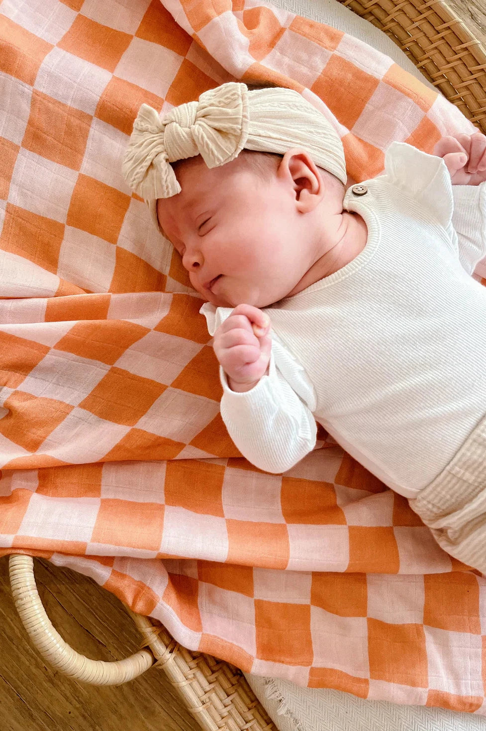 A sleeping baby dressed in a white outfit with a beige bow headband lies on an orange and white checkered blanket from MILKY DESIGNS. The baby is resting in a wicker basket on a wooden floor, wrapped in the buttery softness of the Apricot Peaches Wrap.