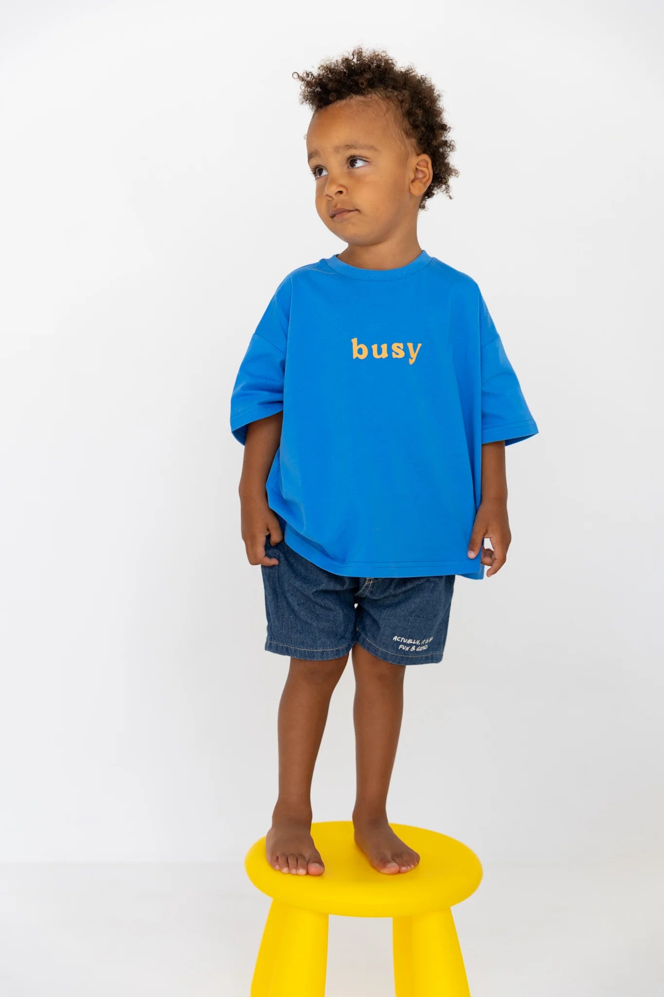 A young child with curly hair stands barefoot on a yellow stool, wearing the LENN LABEL Busy Tee Sunny—an oversized blue kids tee in soft cotton jersey with “busy” printed on it—and dark denim shorts, gazing thoughtfully to the side.