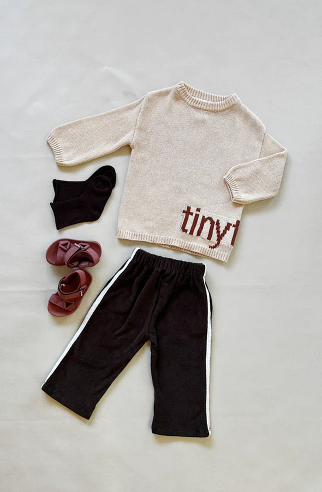 An outfit consisting of a beige sweater with the word "tiny" partially visible on it, TINY TROVE Darcy Racer Pants in dark chocolate featuring an elastic waistband and white side stripes, black socks, and a pair of maroon shoes, all laid out on a light gray surface.