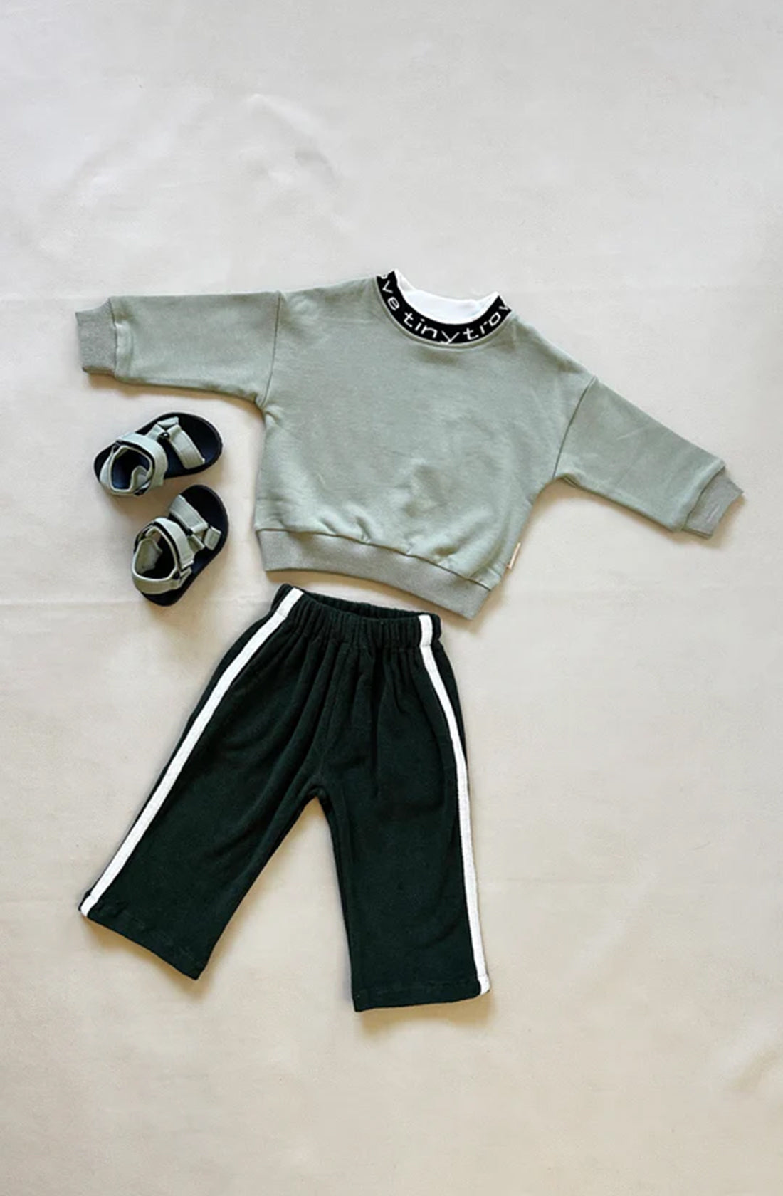 A toddler outfit is laid out on a flat surface. It features a light green long-sleeve shirt with a black collar, Darcy Racer Pants Moss by TINY TROVE which are dark green with an elastic waistband and white side stripes, and a pair of matching green and black shoes. The ensemble ensures comfort and style while being true to size.