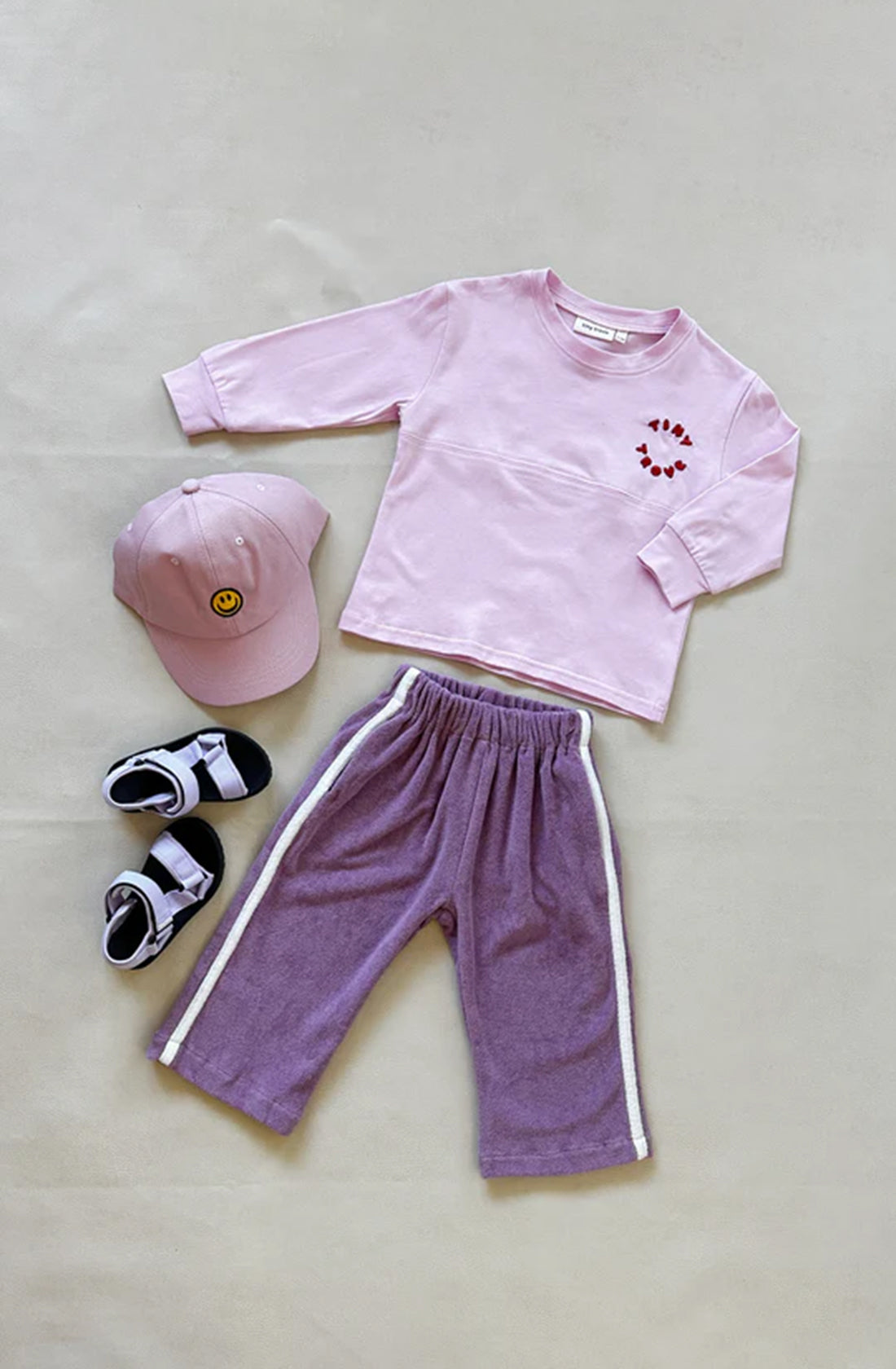 A light pink long-sleeve shirt with tiny red heart decals, paired with loose-fitting Darcy Racer Pants Wisteria from TINY TROVE featuring an elastic waistband, lies on a neutral background. Completing the true-to-size outfit are a pink baseball cap with a smiling face emblem and a pair of black and white sandals.