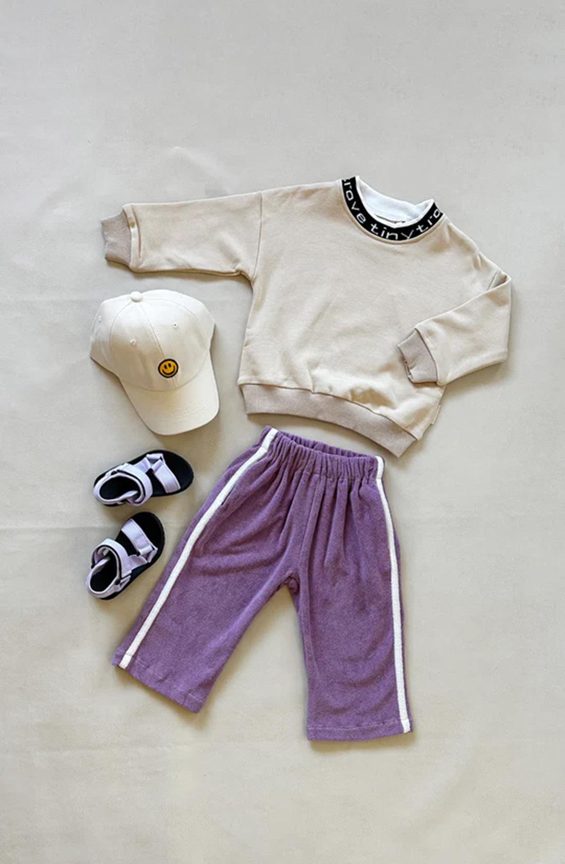 A beige sweatshirt with a black and white collar, paired with TINY TROVE's Darcy Racer Pants in Wisteria featuring white stripes on the sides and an elastic waistband, a beige cap adorned with a yellow smiley face, and white and black sandals are all laid out on a light-colored surface.