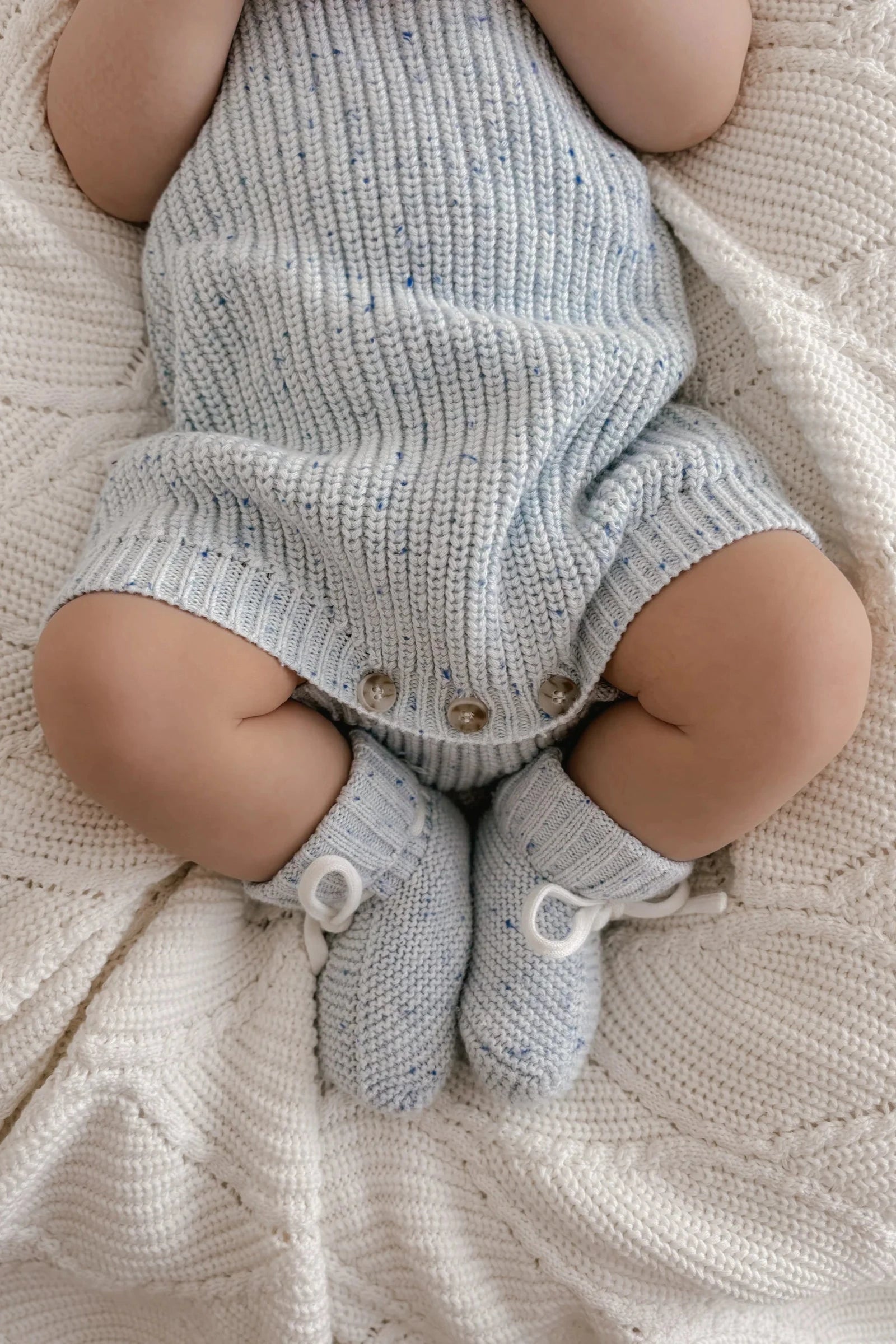 A baby wearing a light blue knit onesie with wooden buttons is lying on a cream-colored knitted blanket. The baby's hands and face are not visible in the image. The scene is completed with ZIGGY LOU's Booties Aero Fleck in matching light blue, adding an adorable touch to the cozy setup.