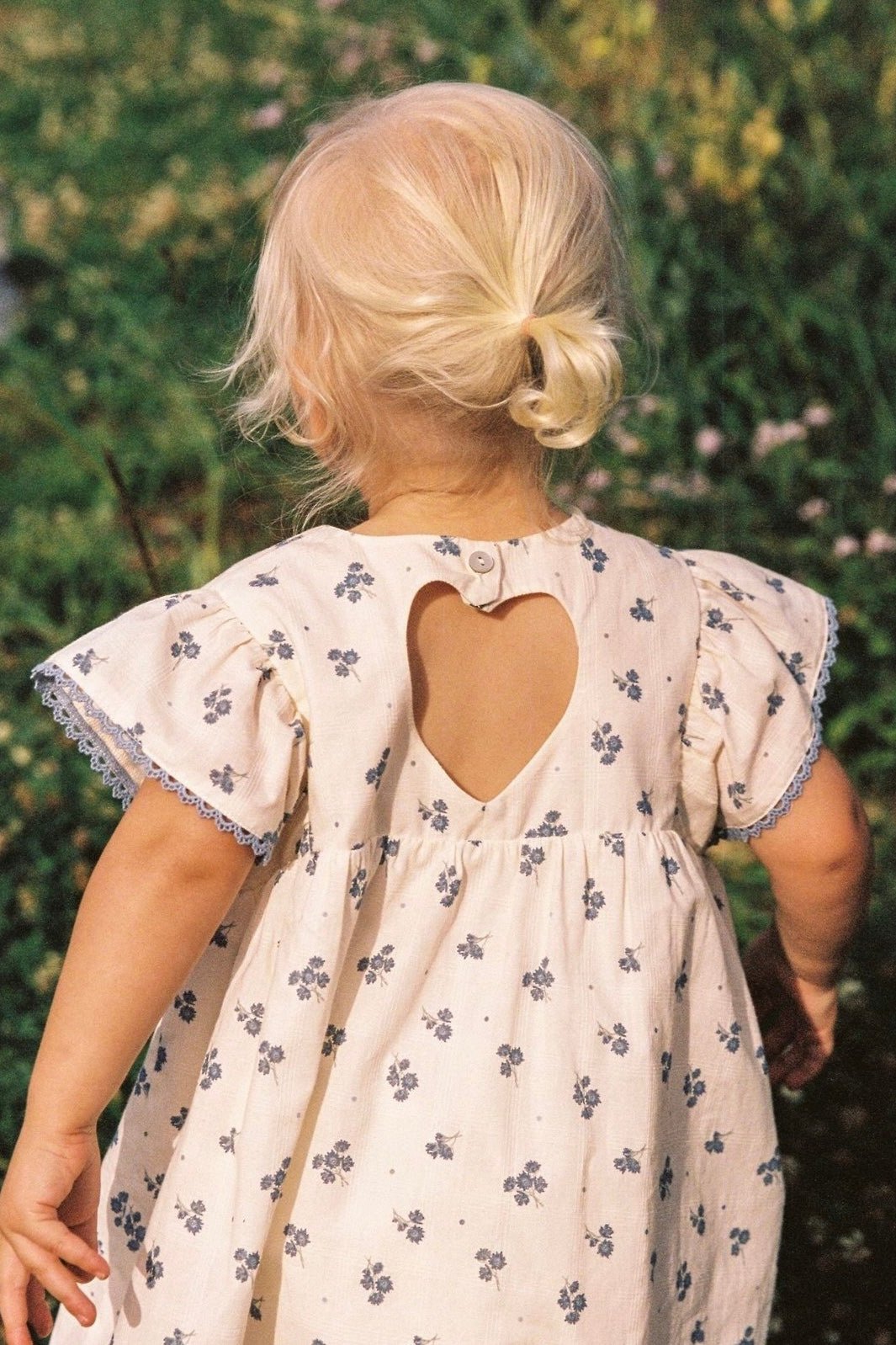 A young child with blonde hair in a small bun wears the Daisy Dress Ditsy Floral from JUNI JUNIOR, featuring delicate lace trim detail and a heart-shaped cutout on the back. The child is standing outdoors, surrounded by greenery and flowers. The child's face is not visible.