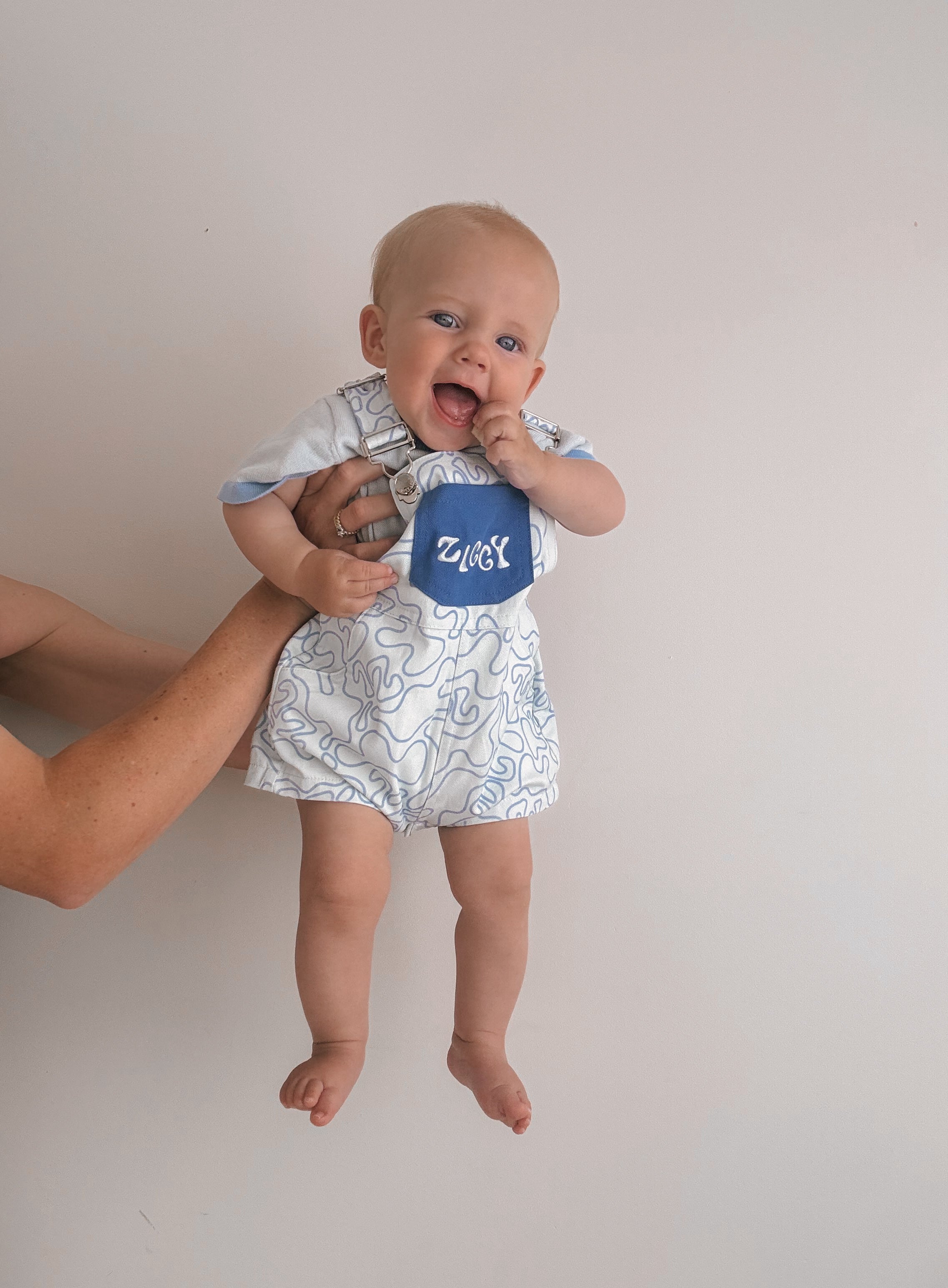 A baby dressed in Short Overalls Zen by ZIGGY LOU, featuring a blue and white pattern and "Ziggy" on the bib, is held up against a plain wall. The baby, with one hand in its mouth and an adorable smile, is supported by an adult's hands from behind.