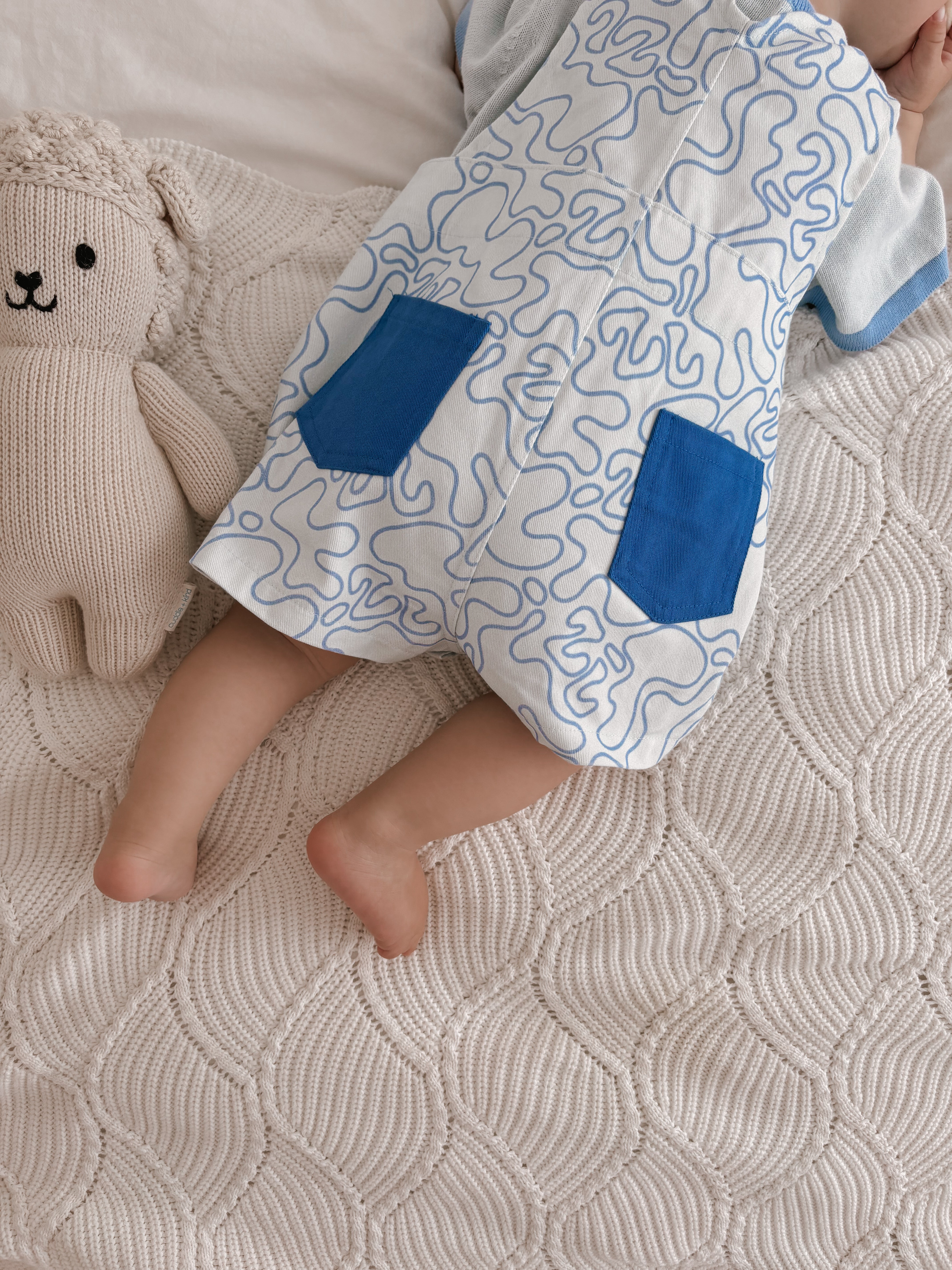 A baby dressed in the ZIGGY LOU Short Overalls Zen, featuring a blue and white pattern, is lying on a textured quilt. The hand-printed design adds charm as their legs stretch out beside a knitted teddy bear, creating a serene and cozy atmosphere.