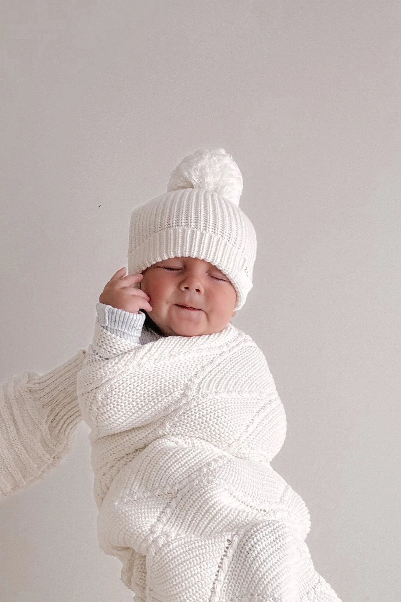 A baby in a cozy white knit outfit and the Ziggy Lou - Beanie Milk by ZIGGY LOU smiles with eyes closed, posing against a plain light-colored background.
