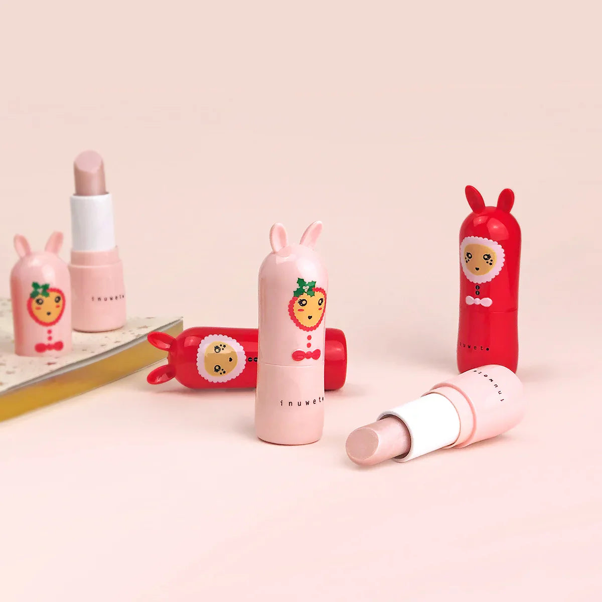 Four INUWET Christmas Lip Balm Candy Cane tubes with playful, animal-themed packaging sit on a light surface. Two tubes are opened, revealing light pink balm made with natural ingredients. The colorful illustrations resemble a rabbit and a lion, adorned with bows and fruit motifs, highlighting their cruelty-free appeal.