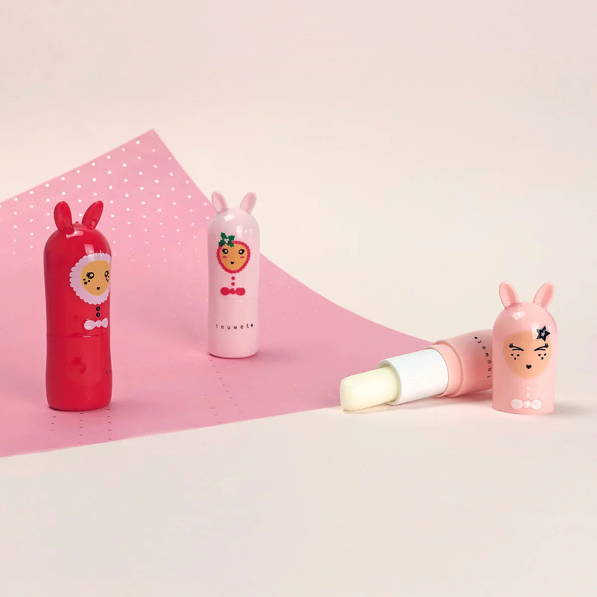 Three INUWET lip balms featuring adorable character designs: one red with a smiling face, one light pink with an open cap revealing the cruelty-free balm, and one peach with a strawberry face. Each is topped with bunny ears against a pink and white background, made from natural ingredients.