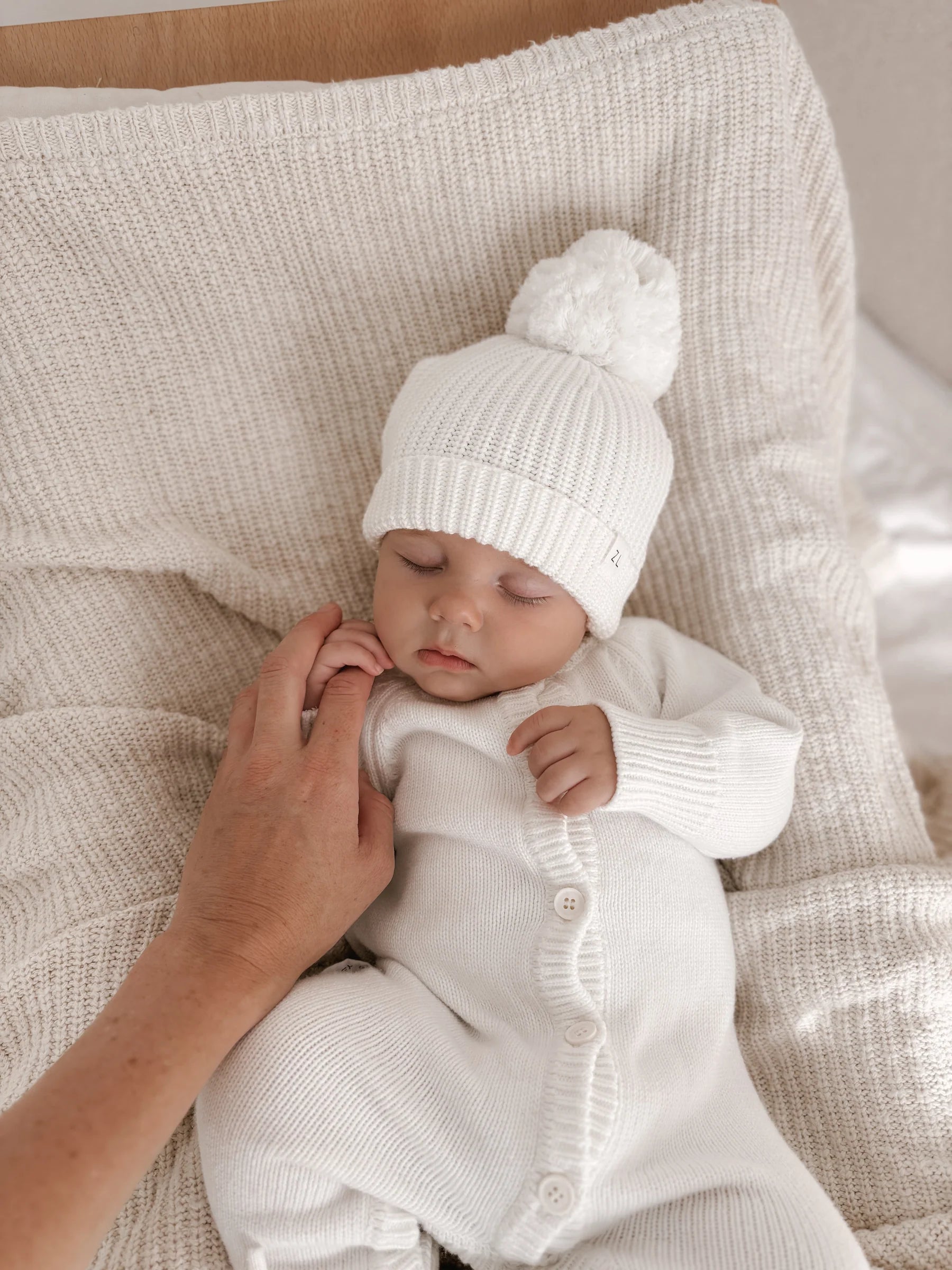 A baby wearing the Ziggy Lou - Classic Knit Romper Milk by ZIGGY LOU sleeps on a cream blanket as an adult gently touches their hand, highlighting this cozy winter outfit from ZIGGY LOU.