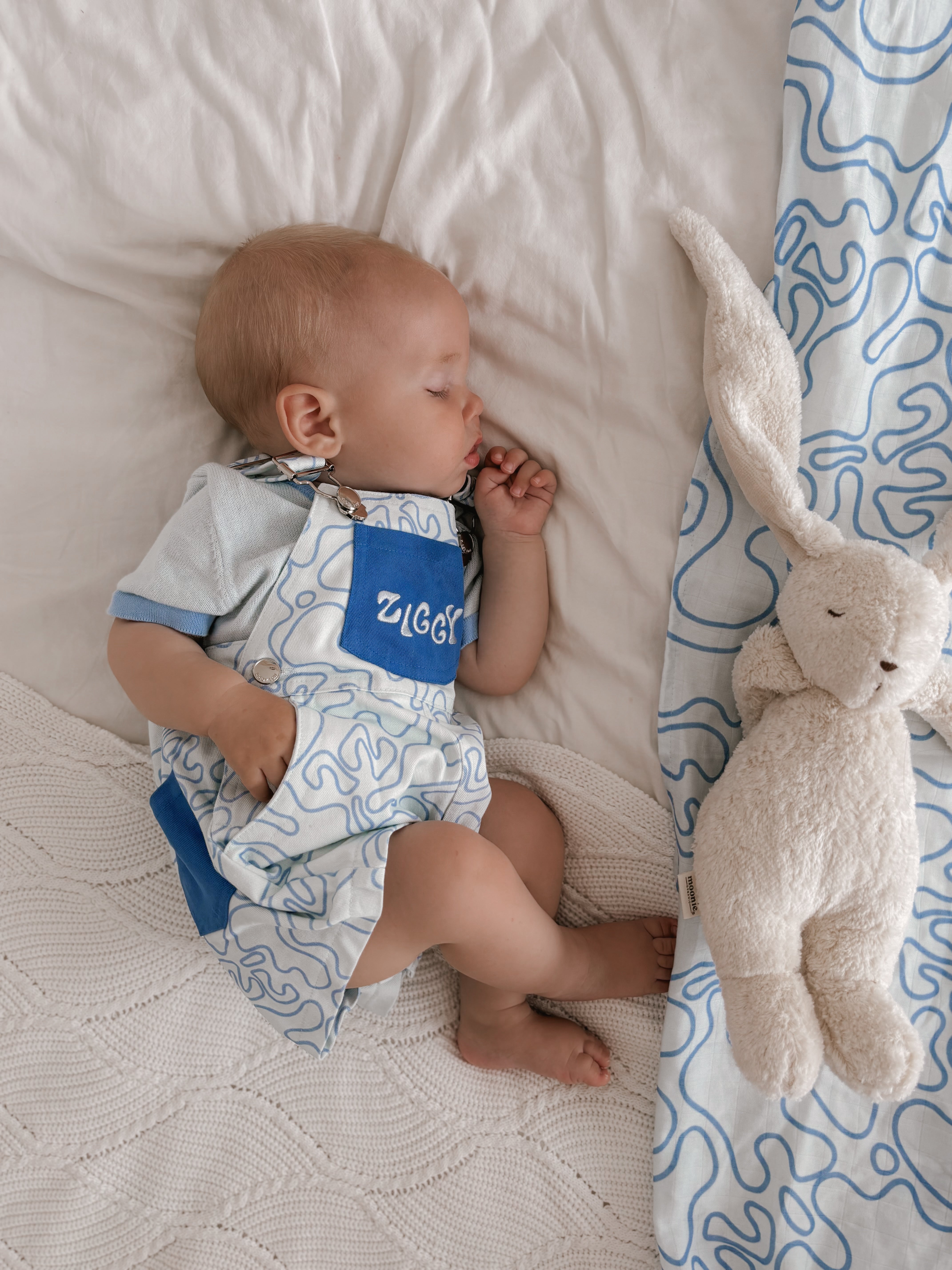 A baby sleeps on a white blanket, dressed in blue and white Short Overalls Zen by ZIGGY LOU, with "Ziggy" on the pocket. A plush rabbit toy rests nearby, alongside a patterned blue and white cloth, all crafted from 100% cotton canvas for comfort and style.