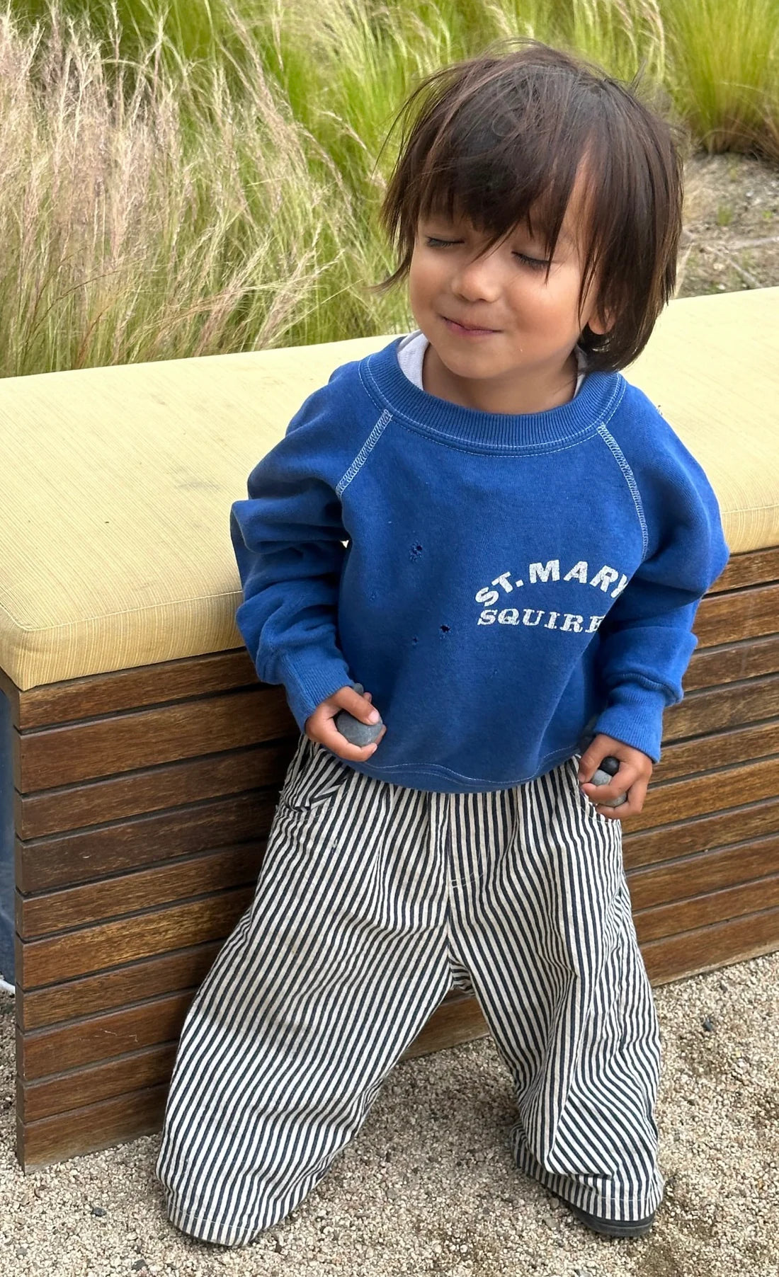 A young child with medium-length brown hair smiles with eyes closed, wearing a blue "St. Mary Squirrels" sweatshirt and ZIGGY ZAZA Ziggy Zaza - Casper Pant Navy Stripe, standing in front of a wooden bench and tall grass.