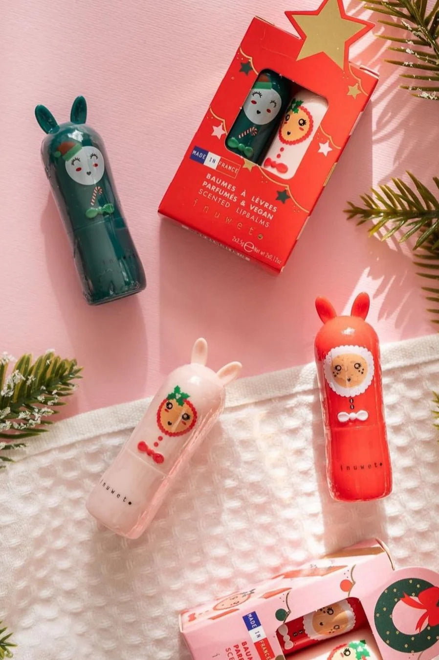 A festive display showcases INUWET's Christmas Lip Balm Cherry, featuring colorful, vegan lip balm tubes shaped like animal figures on a pink background. The arrangement is holiday-themed with decorations such as stars and wreaths. A partially opened box reveals two cherry-scented lip balms, while pine branches enhance the corners.